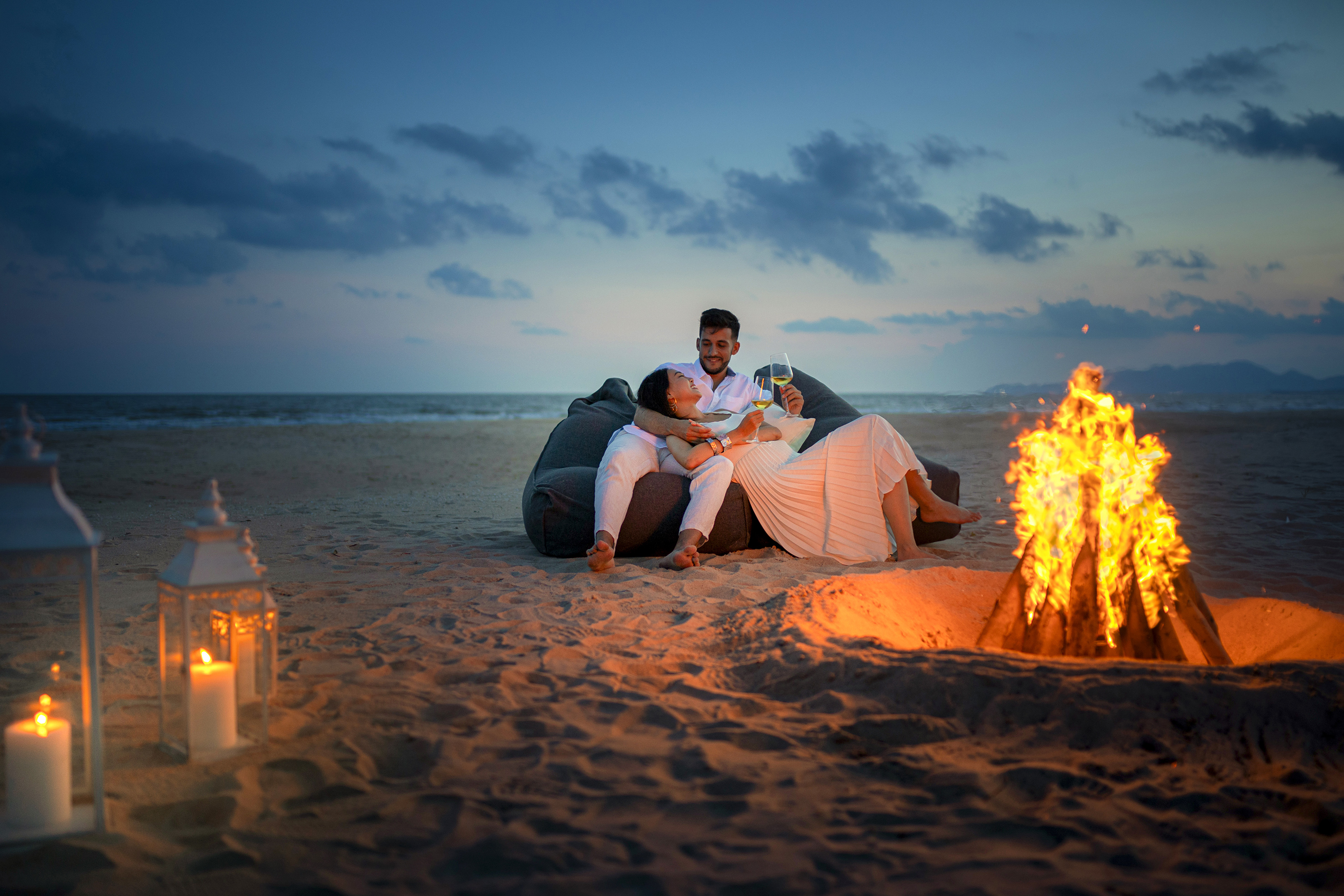 Romantic couple on beach with bonfire, wine, lanterns, and ocean at twilight.
