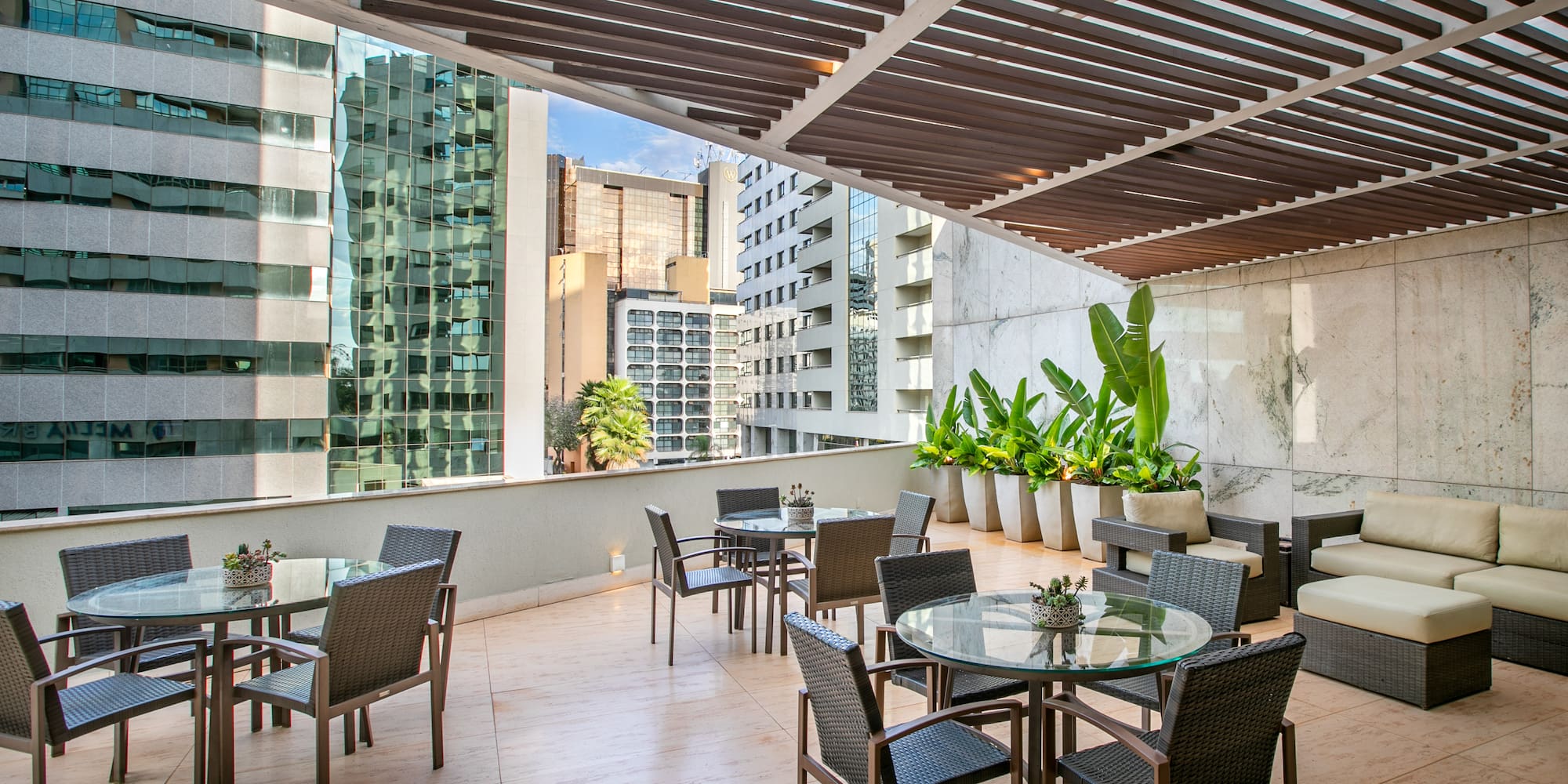 Modern outdoor terrace with dining tables, lounge, green plants, and city skyline.