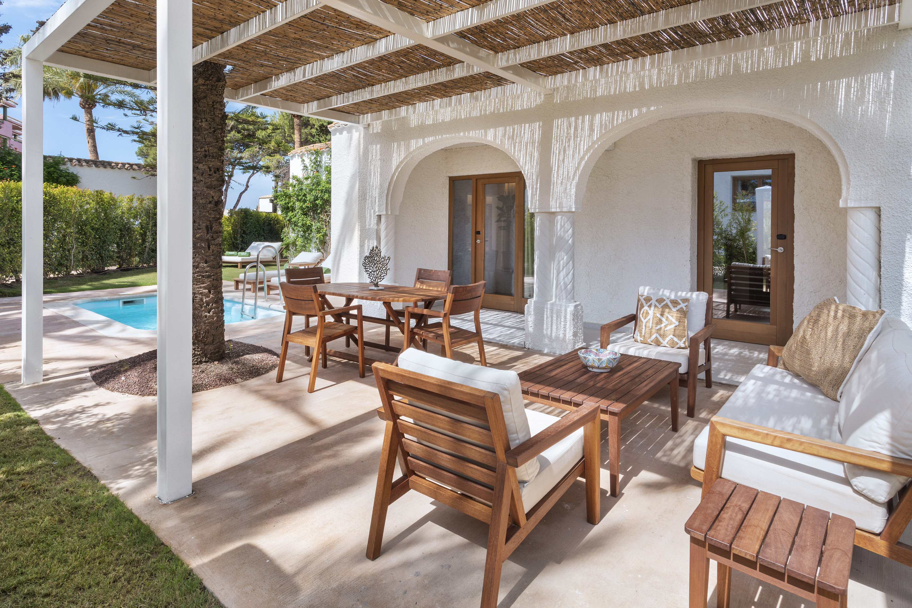 Sunny outdoor patio with wooden furniture, woven pergola, and swimming pool.