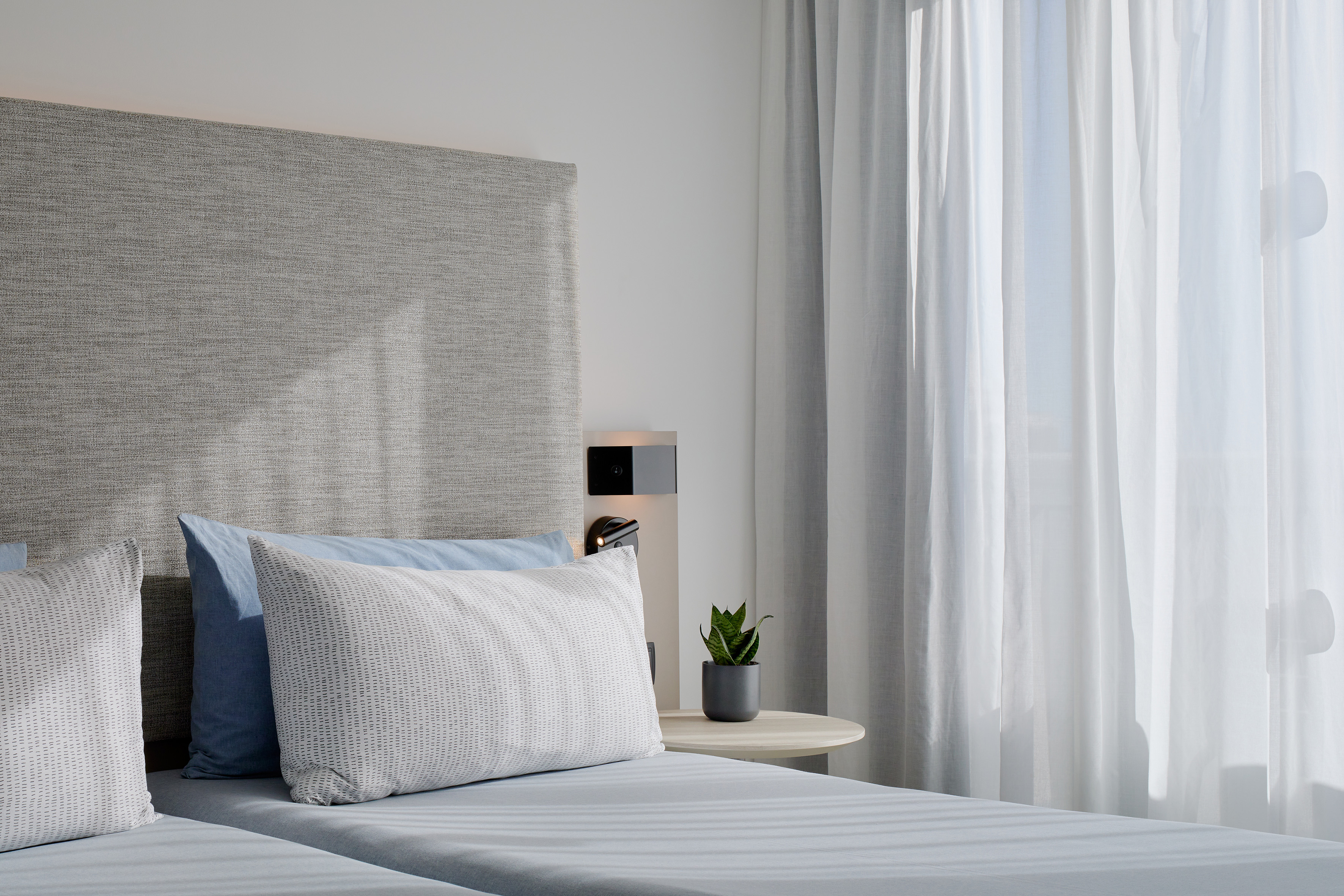 Bright hotel bedroom with blue bed, grey headboard, and sheer window curtains.