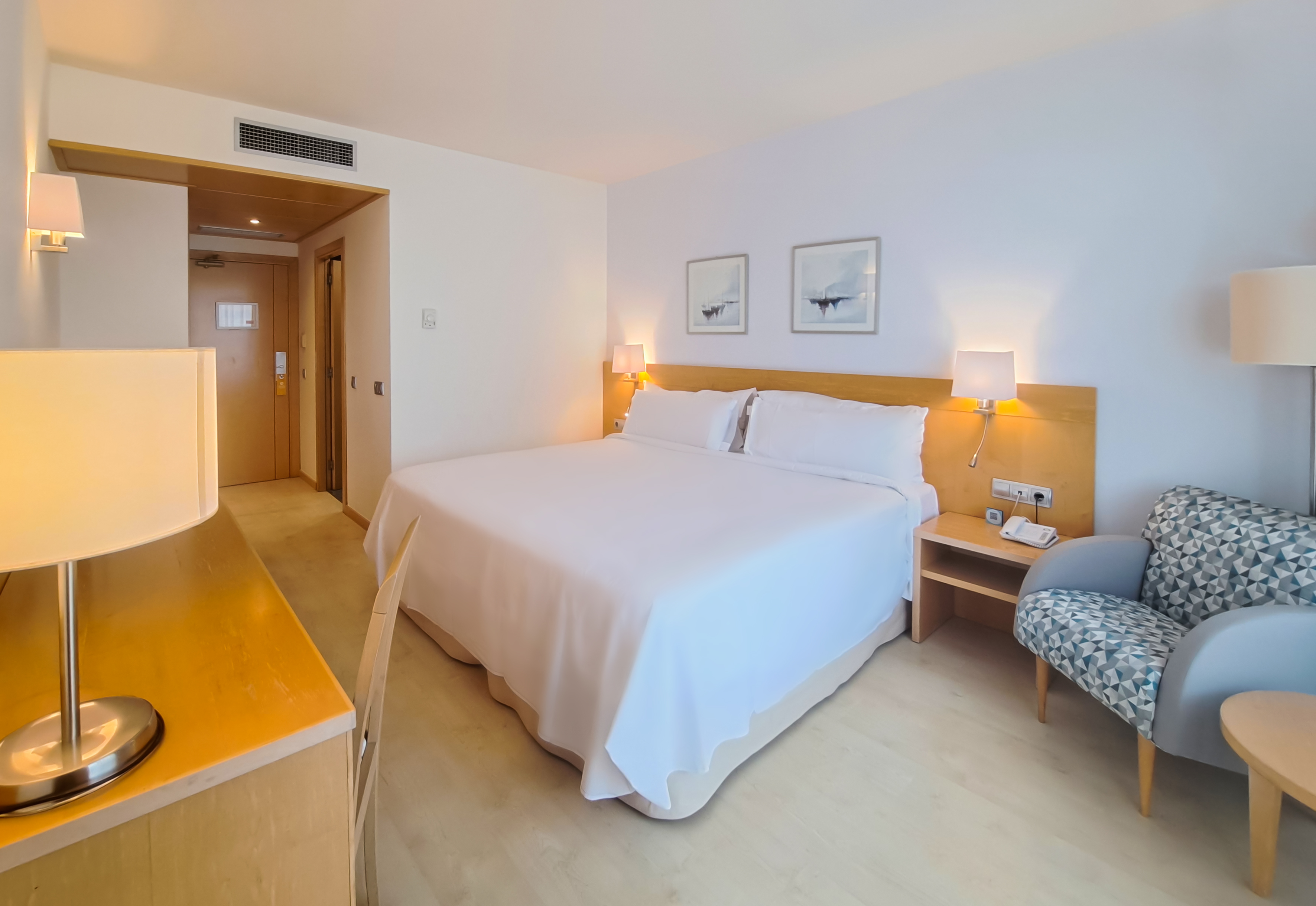 Modern hotel bedroom with large white bed, wooden desk, and patterned armchair.