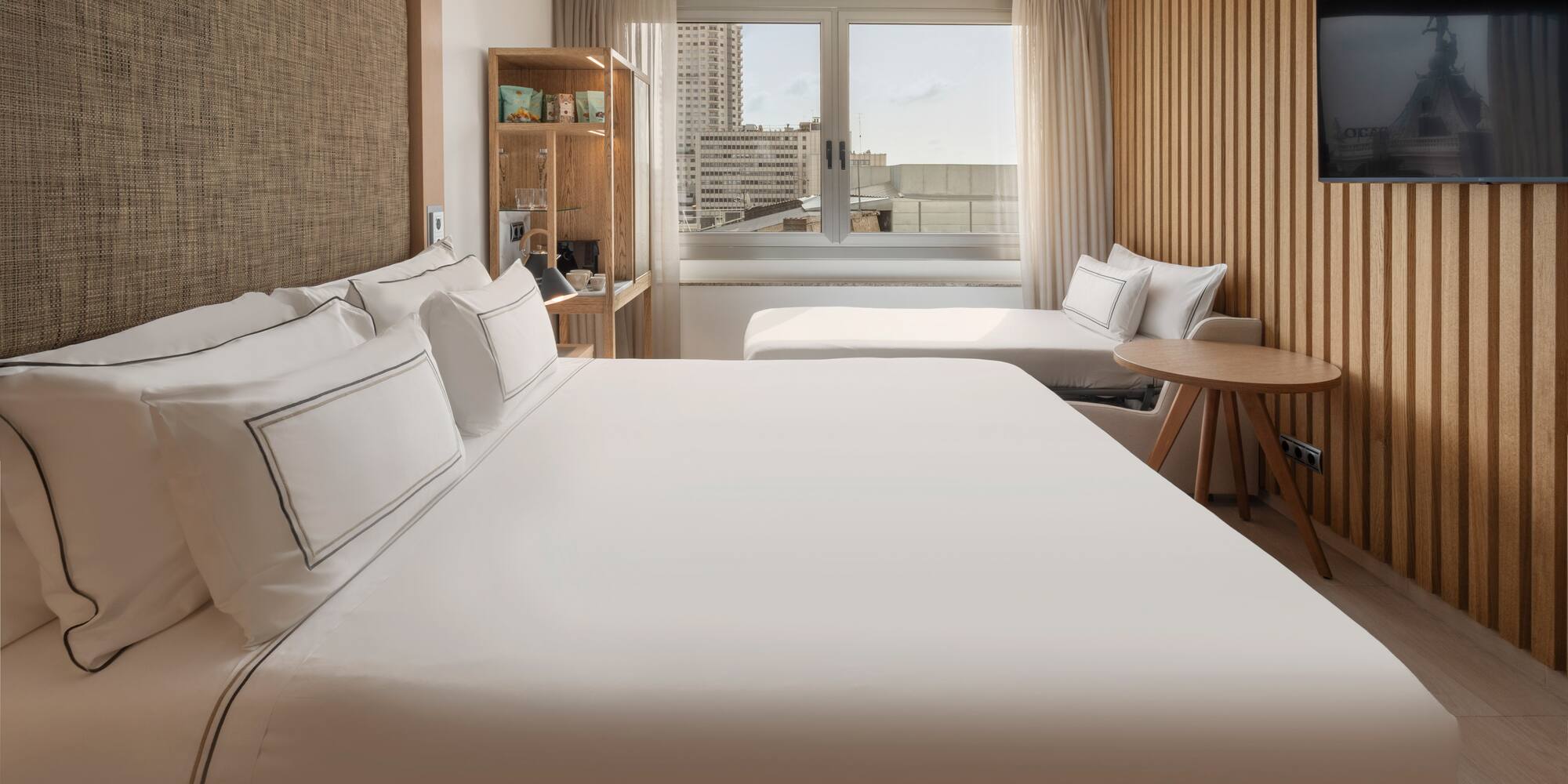 Modern hotel room with large bed, city view, wooden decor, and a daybed.
