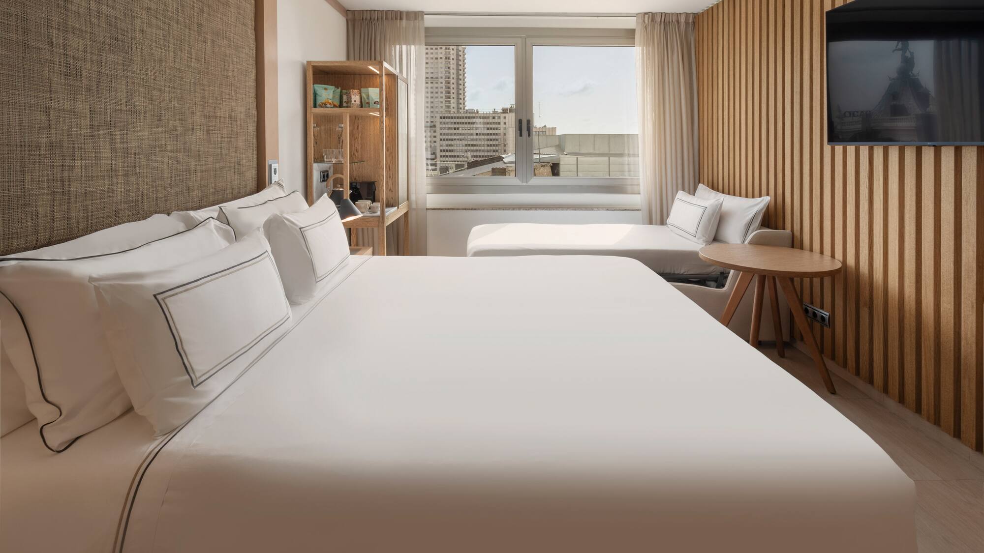 Modern hotel room with large bed, city view, wooden decor, and a daybed.