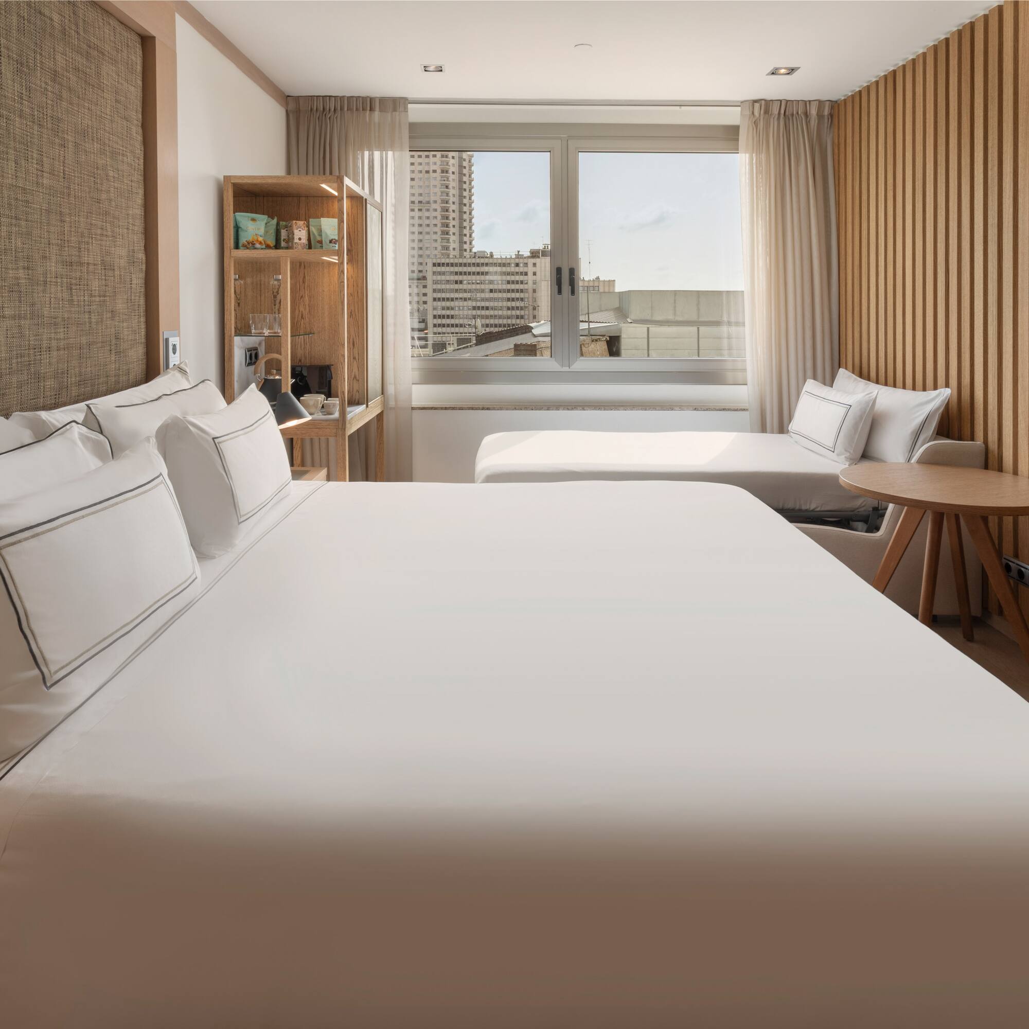Modern hotel room with large bed, city view, wooden decor, and a daybed.