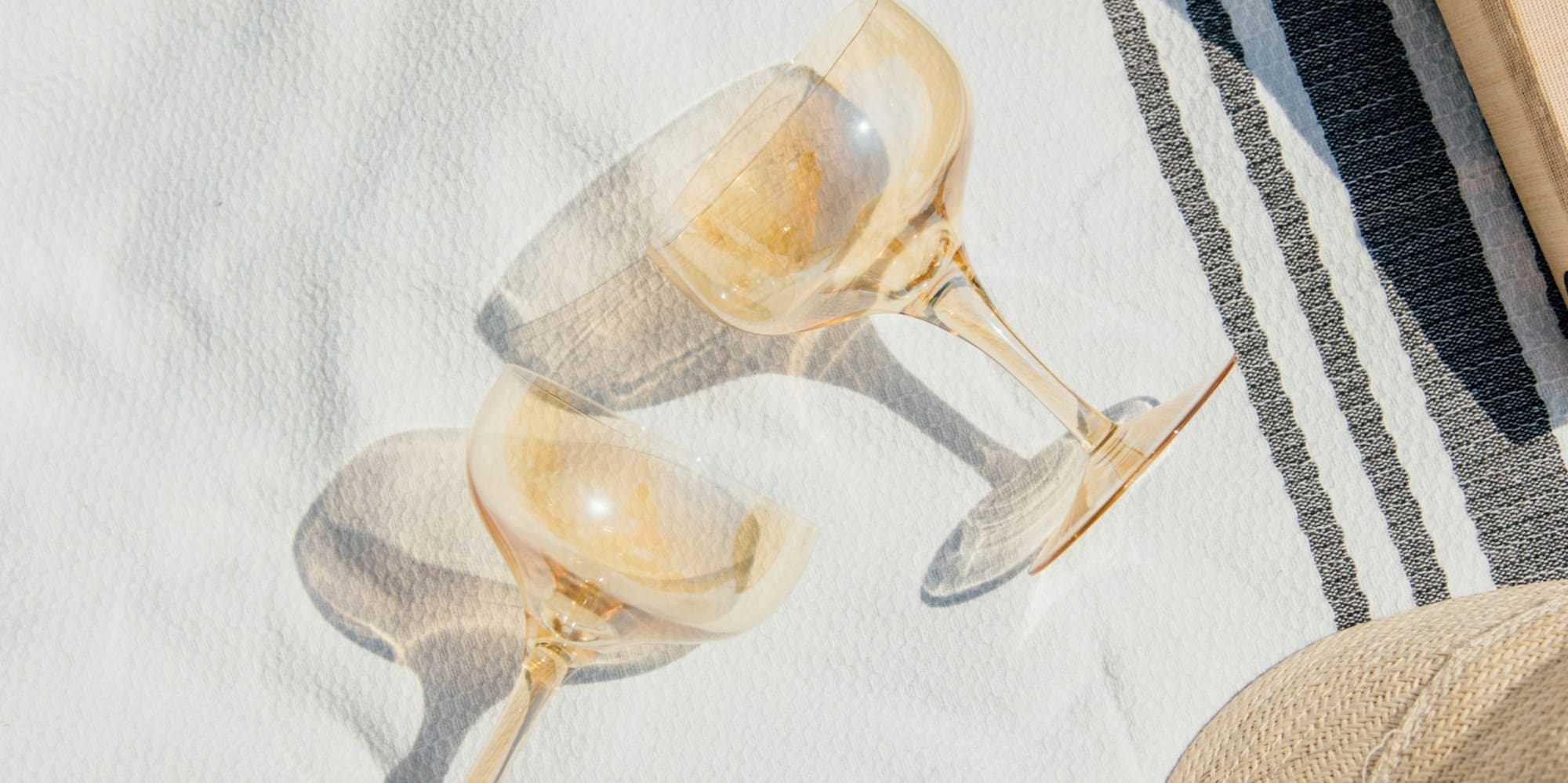 Outdoor picnic: sunscreen, books, champagne glasses, straw hat on white blanket.