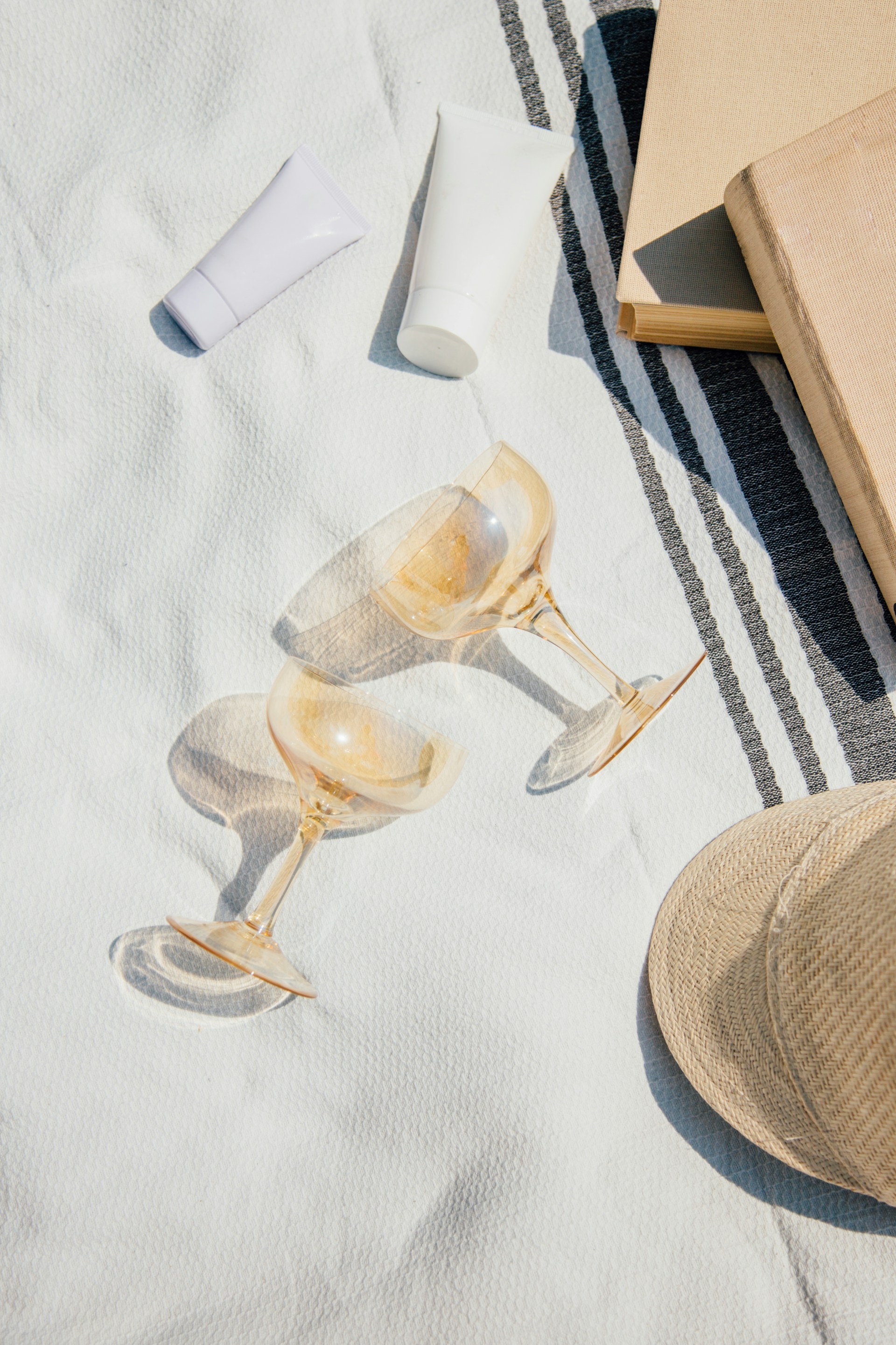Outdoor picnic: sunscreen, books, champagne glasses, straw hat on white blanket.