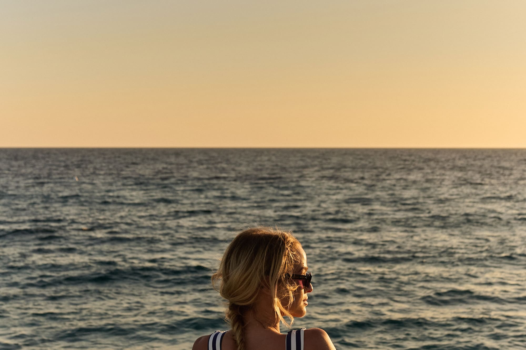 Woman in striped swimsuit on boat, looking at ocean sunset.
