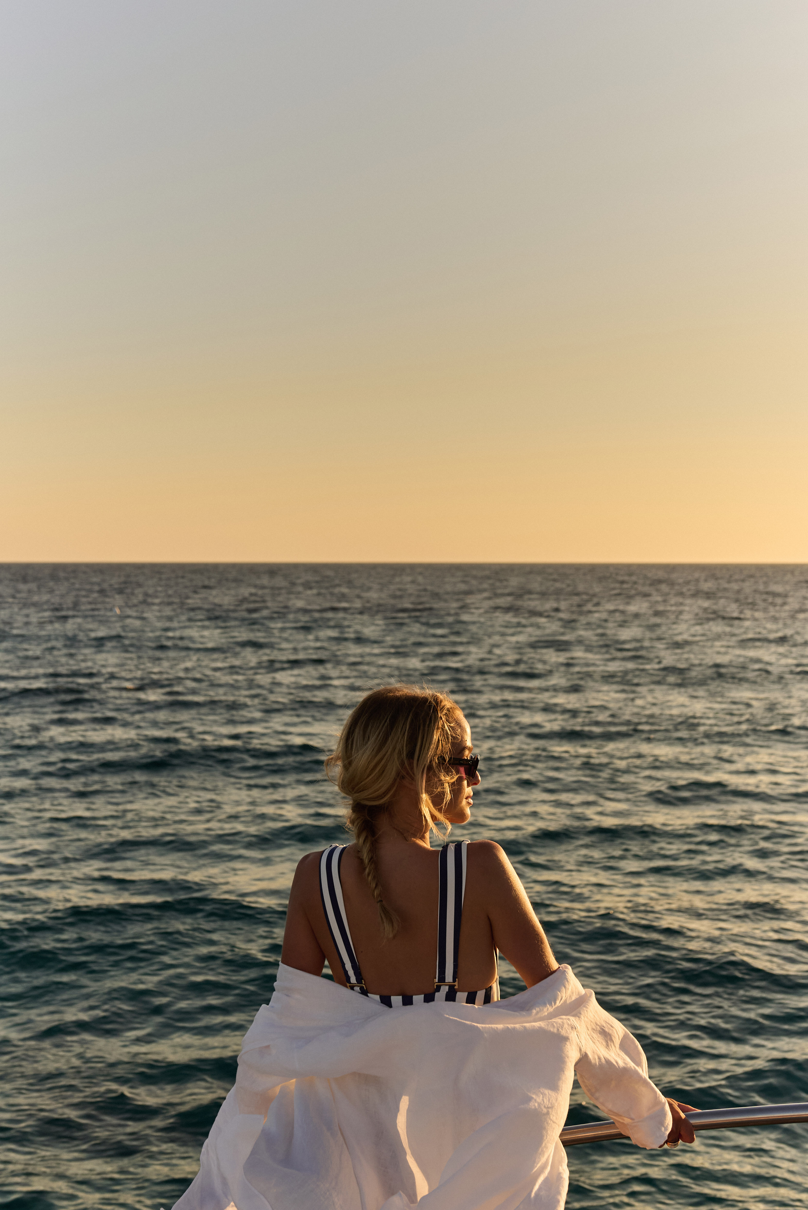 Woman in striped swimsuit on boat, looking at ocean sunset.
