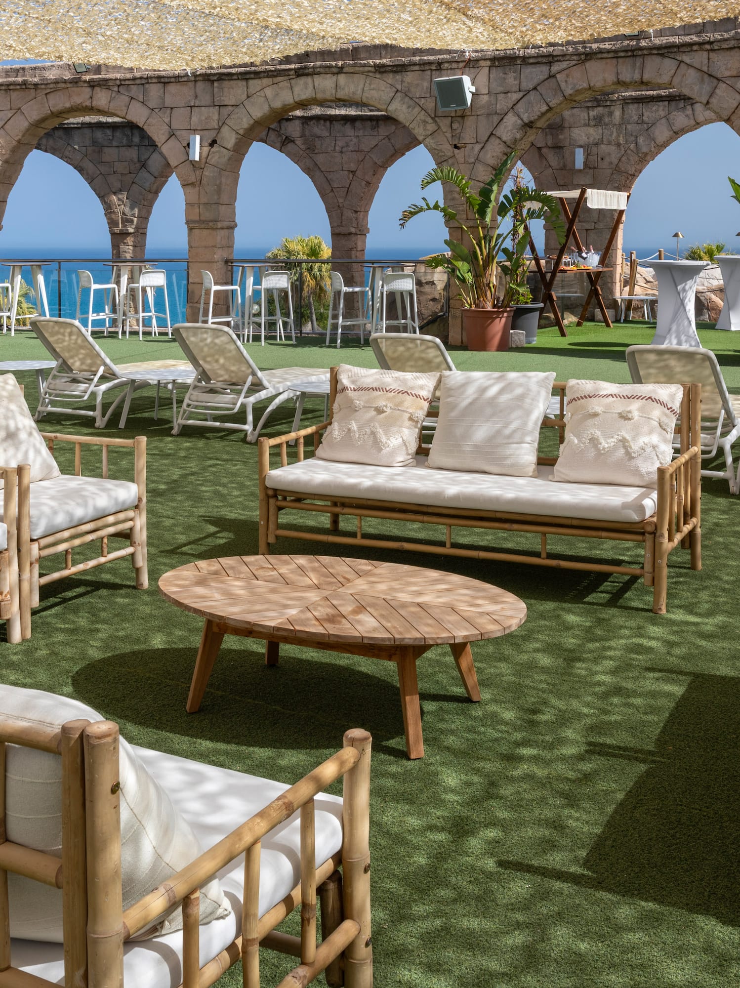 Bamboo lounge seating on artificial grass patio with ocean view and stone arches.