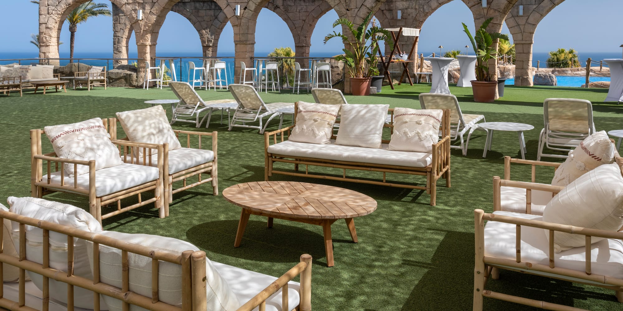 Bamboo lounge furniture on green turf with stone arches and ocean view.