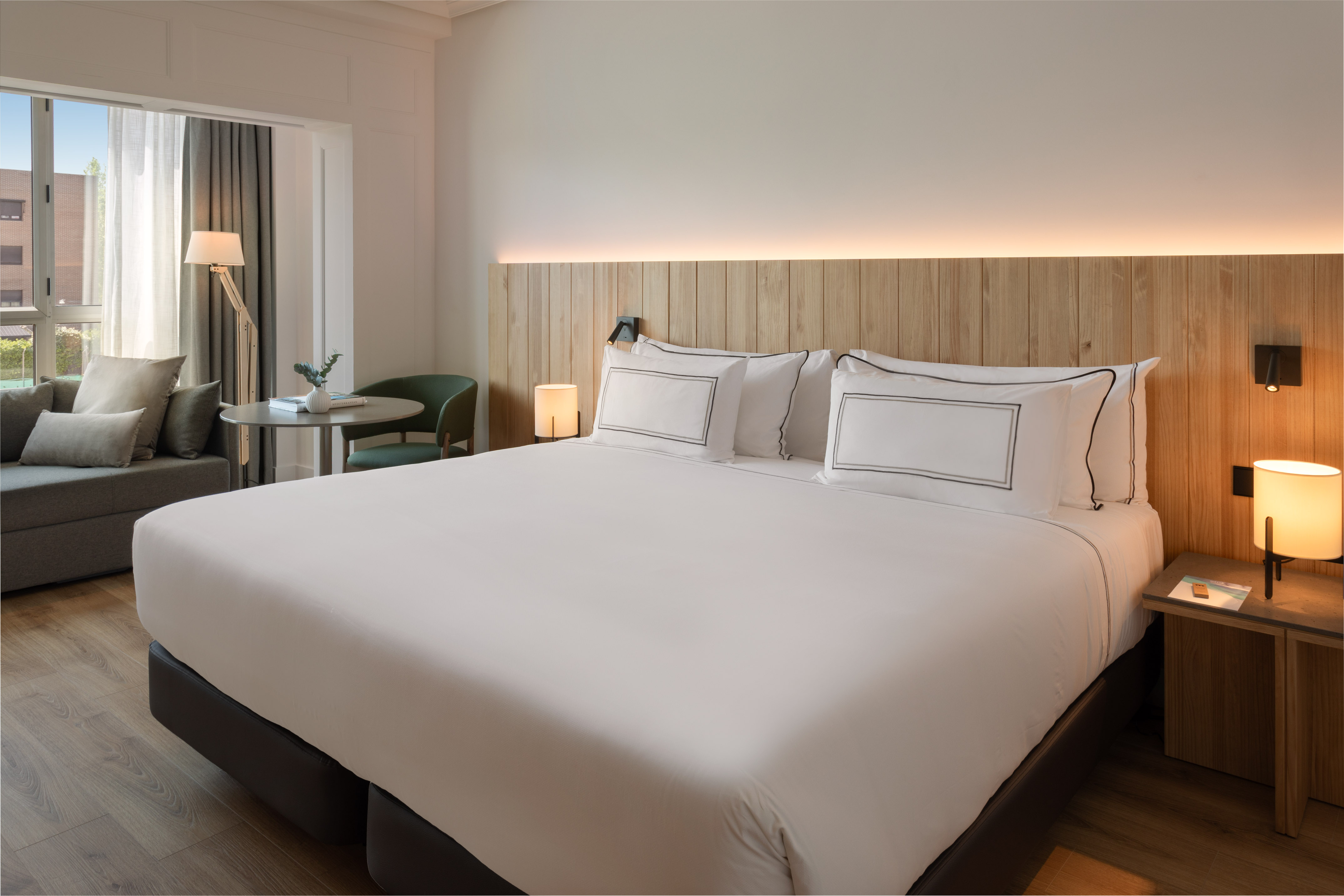 Hotel room with king bed, wooden headboard, and sunlit window-side sitting area.