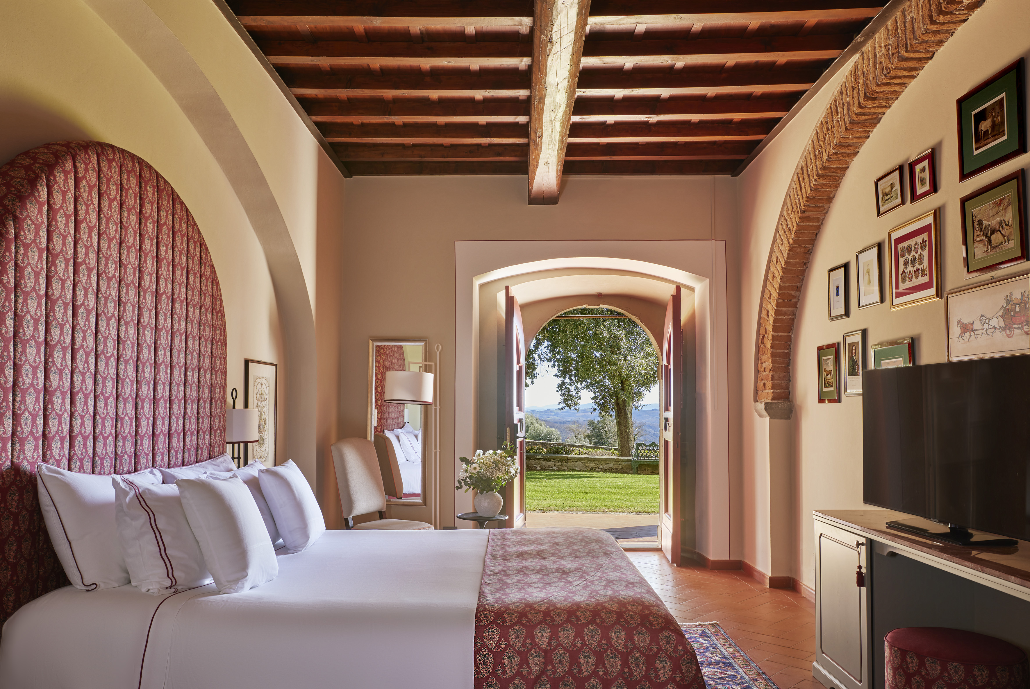 Luxury Tuscan hotel room with red bed, wood beams, and arched garden view.