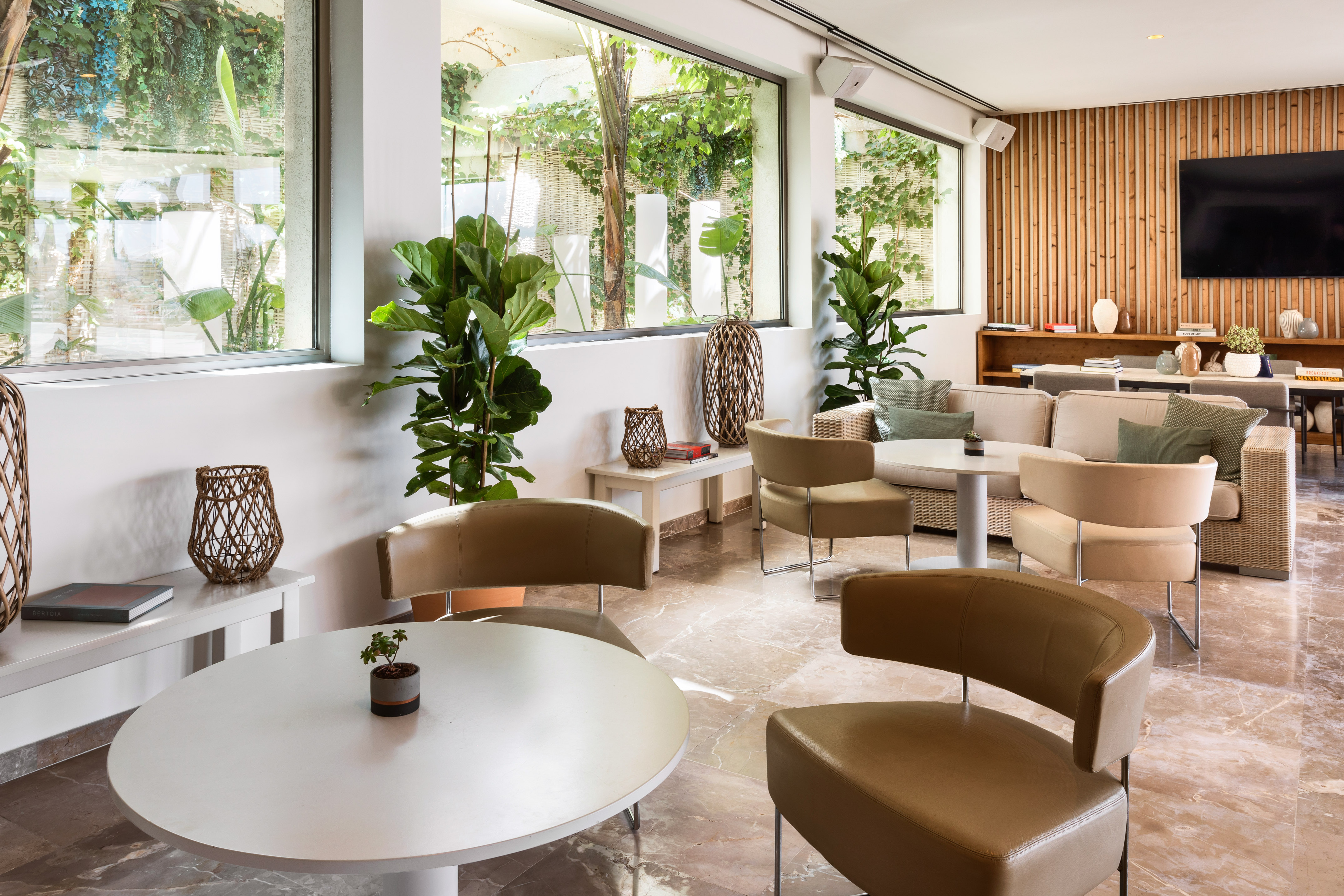 Modern lounge with large windows, indoor plants, marble floor, and comfortable beige seating.