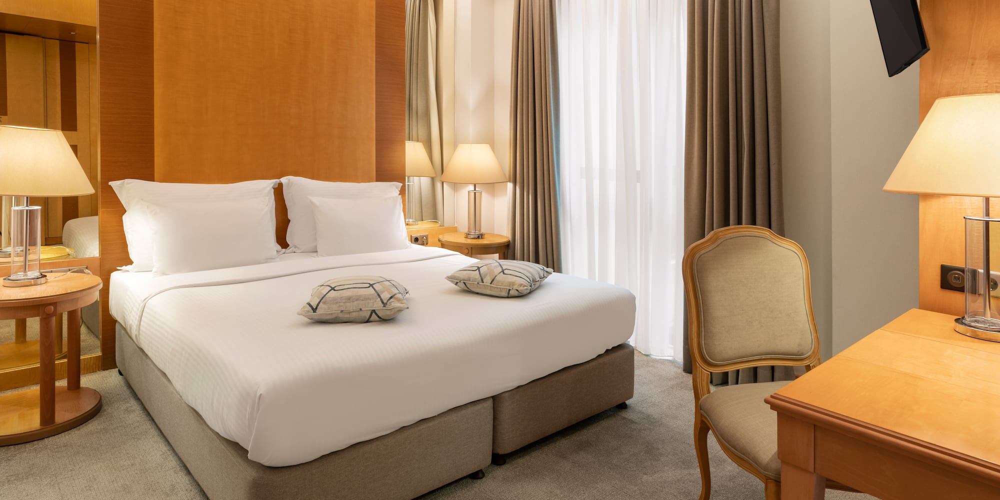 Modern hotel bedroom with king bed, wood headboard, lamps, desk, and TV.