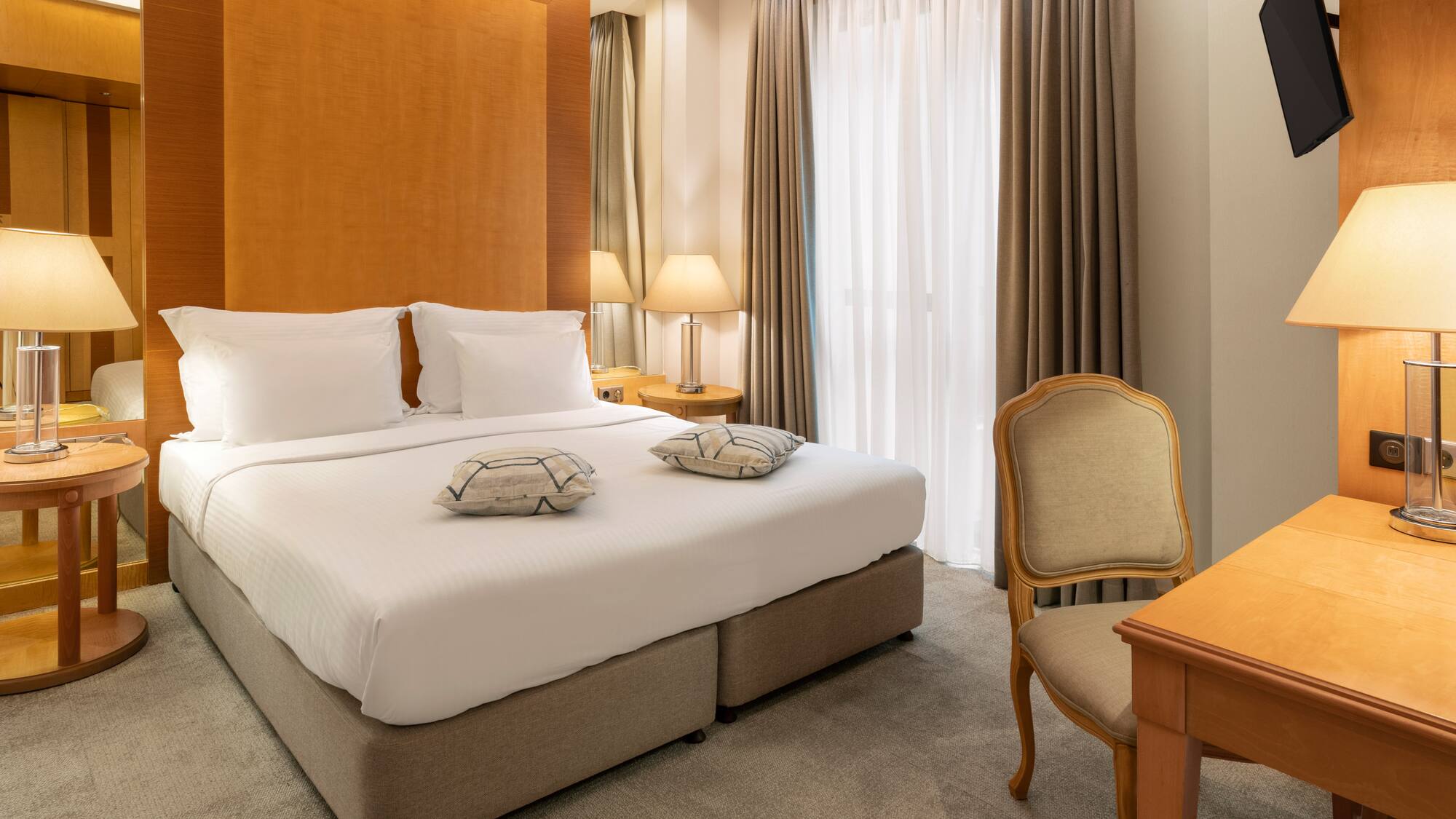 Modern hotel bedroom with king bed, wood headboard, lamps, desk, and TV.