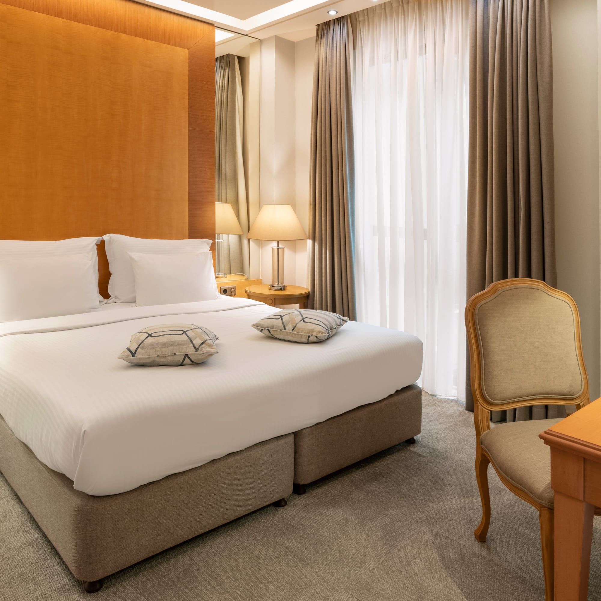 Modern hotel bedroom with king bed, wood headboard, lamps, desk, and TV.
