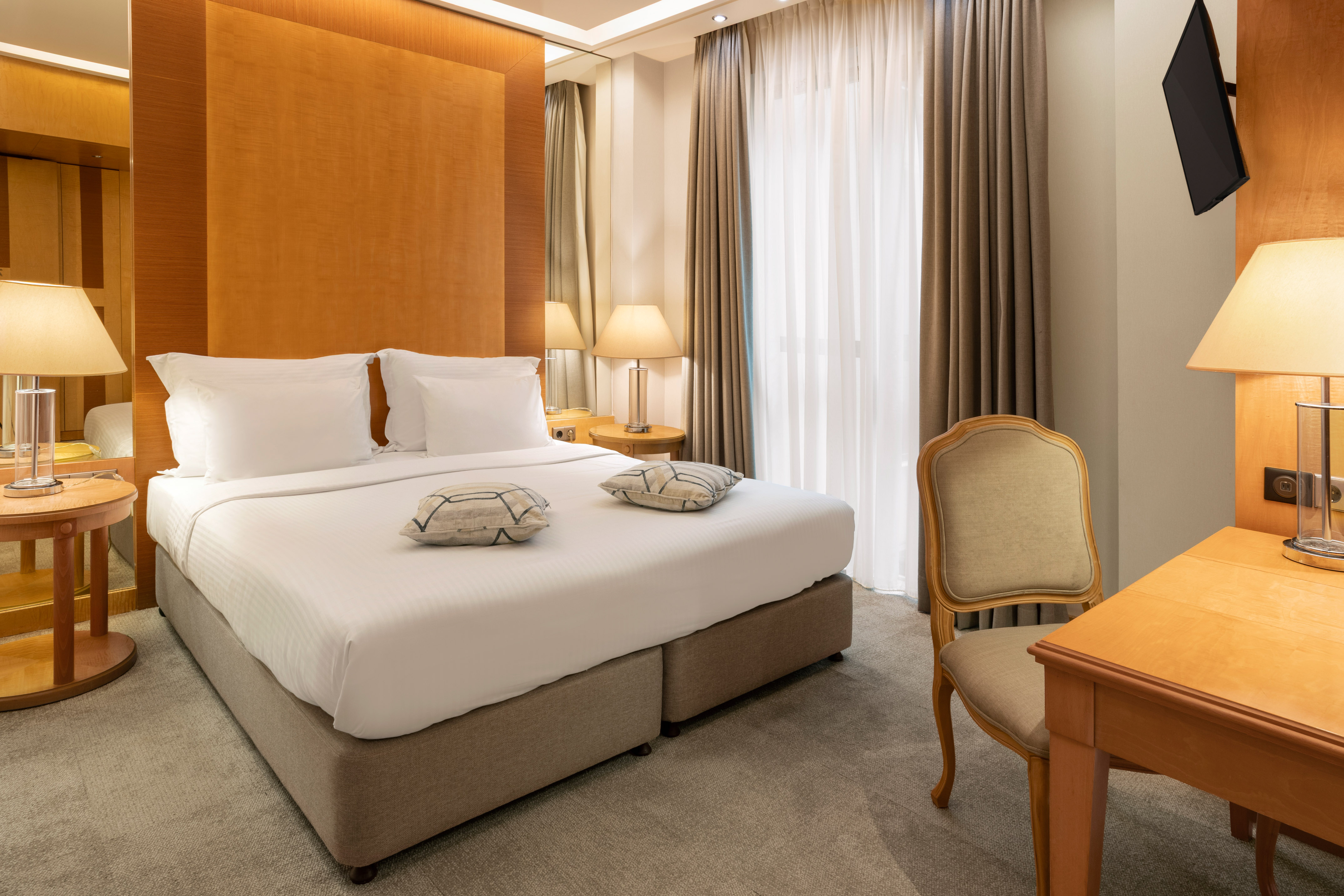Modern hotel bedroom with king bed, wood headboard, lamps, desk, and TV.