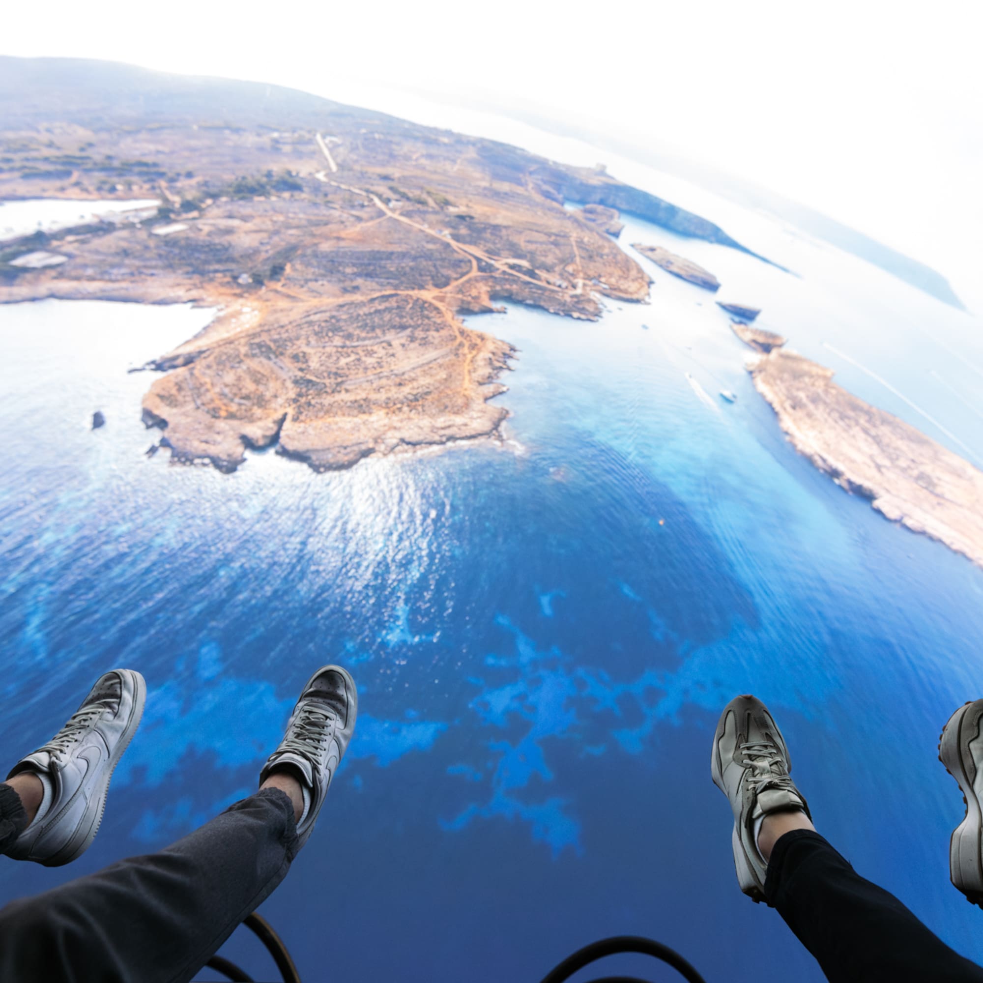 Feet dangling high above vast blue ocean and rugged islands, aerial perspective.