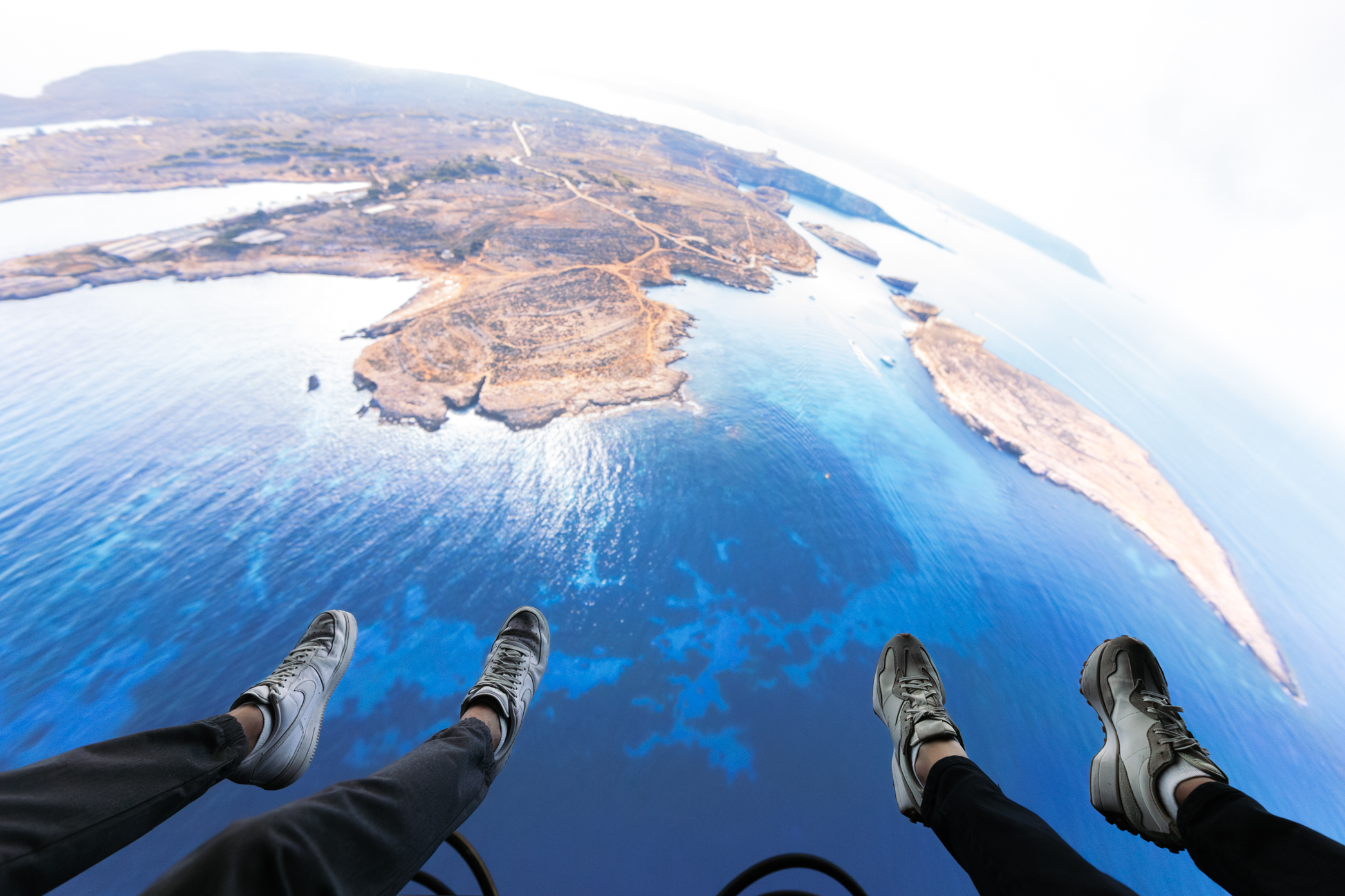 Feet dangling high above vast blue ocean and rugged islands, aerial perspective.