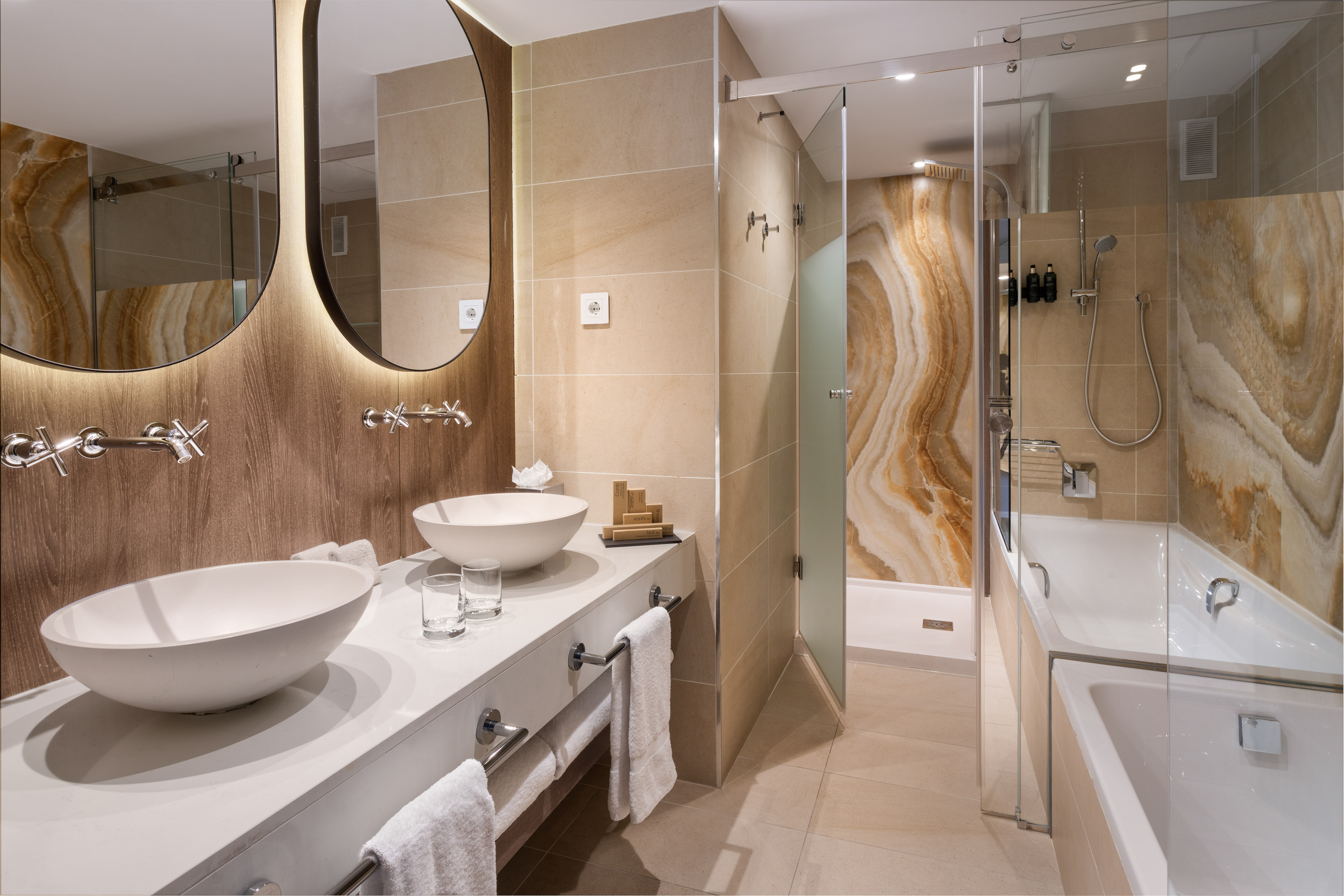 Modern hotel bathroom: dual sinks, backlit mirrors, glass shower, and tub.