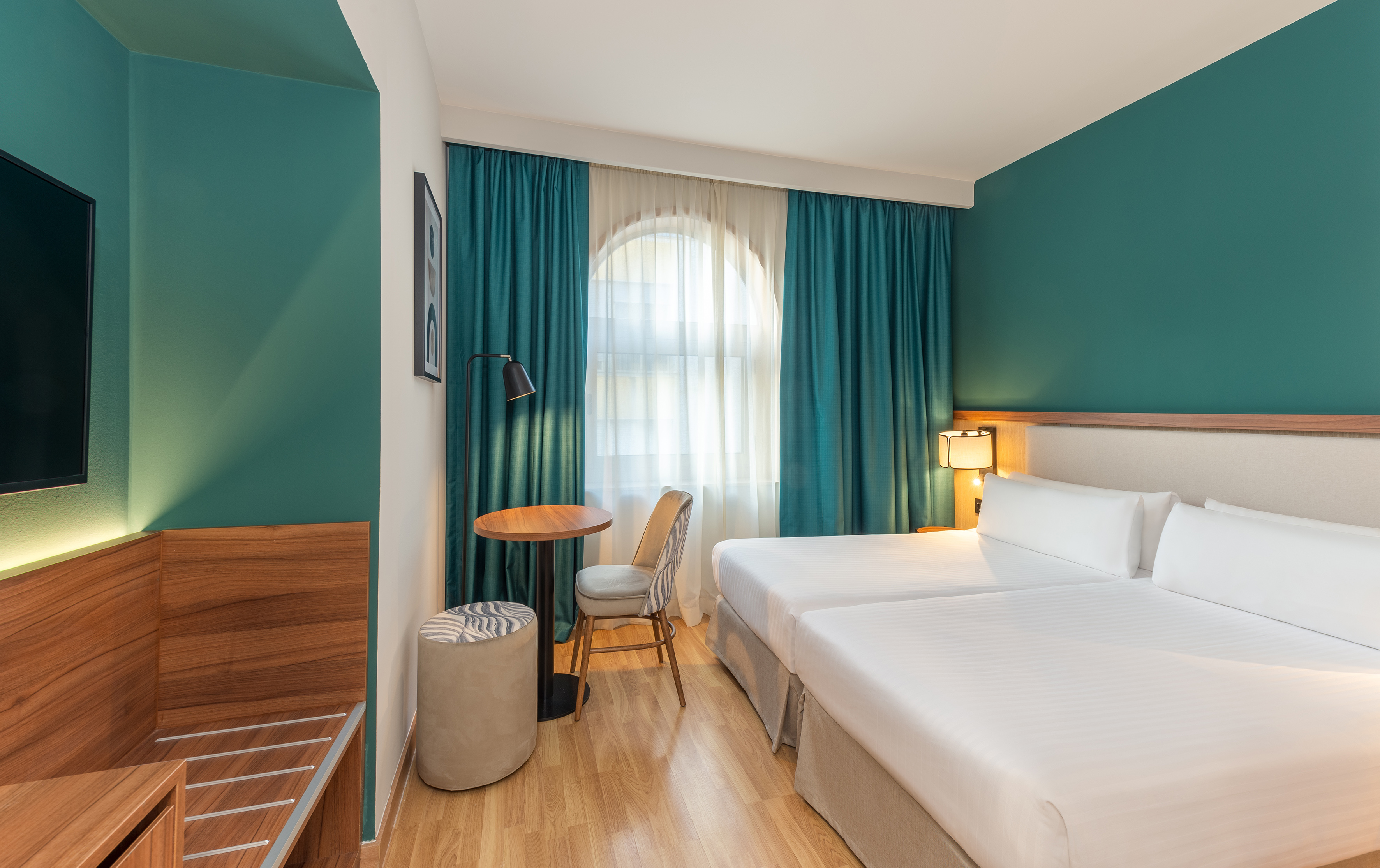 Modern hotel room with twin beds, teal walls, arched window, and wood floor.