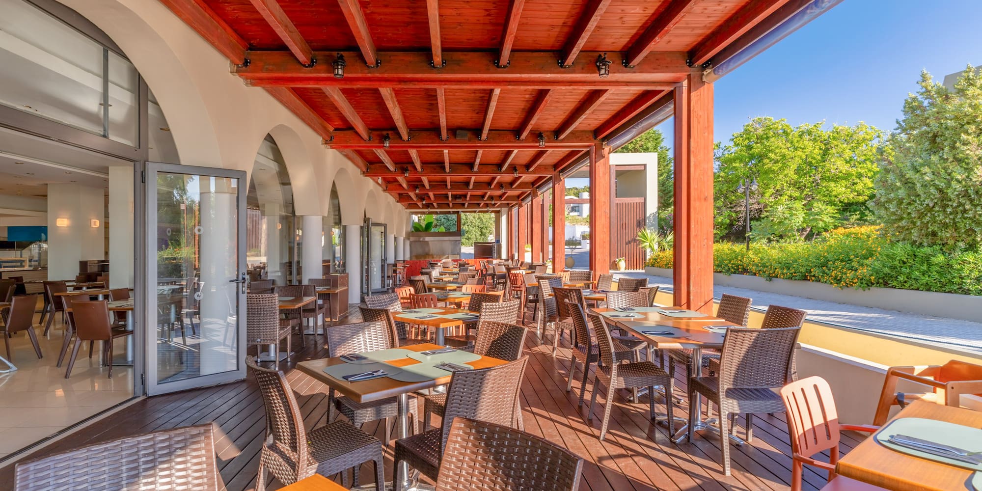 Sunny outdoor restaurant patio with wooden deck, tables, chairs, and red beamed roof.