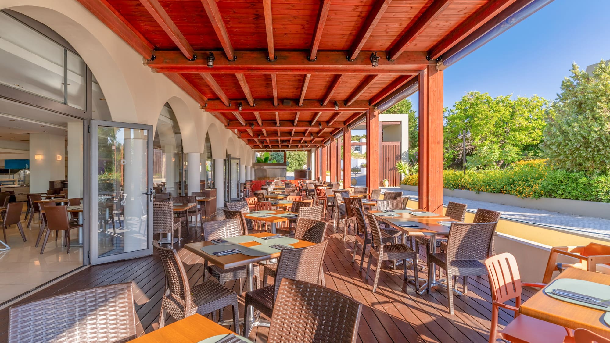 Sunny outdoor restaurant patio with wooden deck, tables, chairs, and red beamed roof.