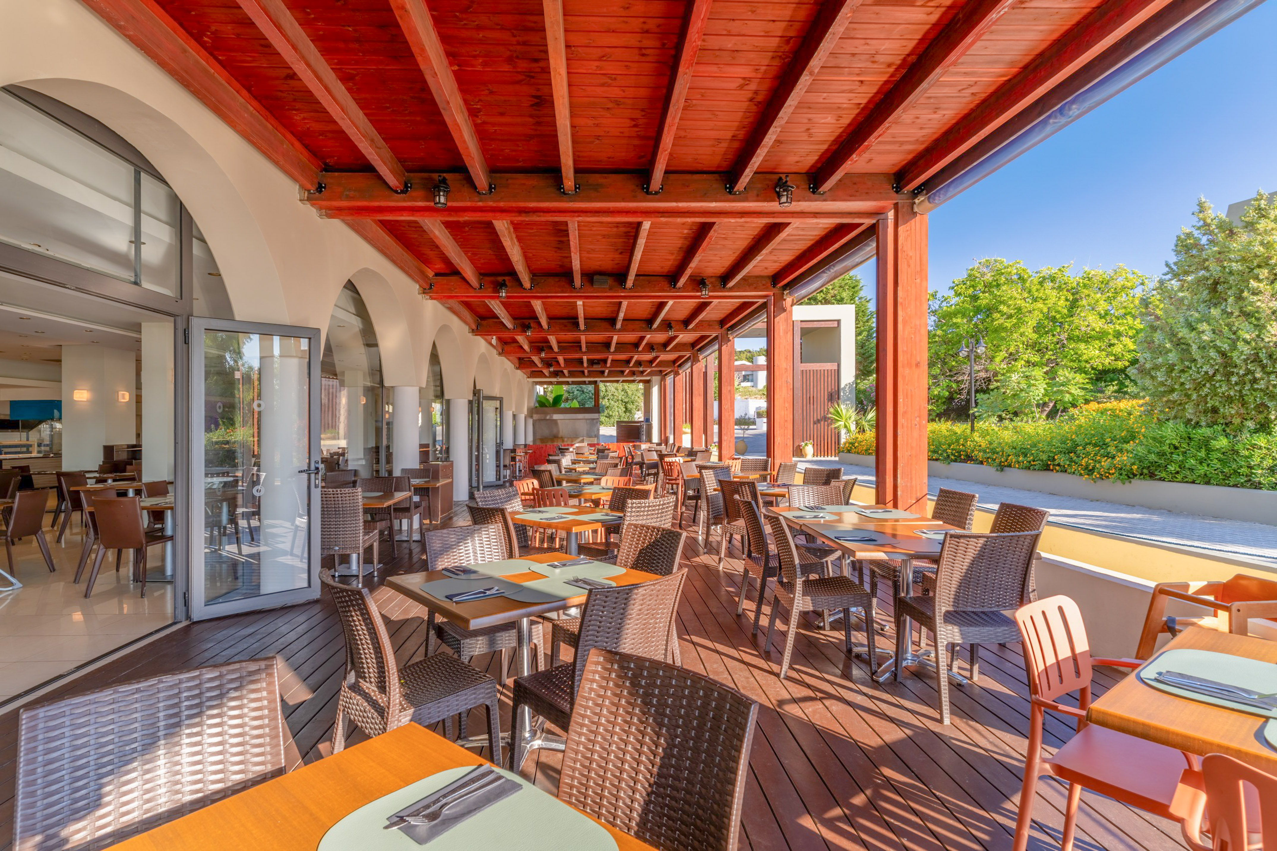 Sunny outdoor restaurant patio with wooden deck, tables, chairs, and red beamed roof.