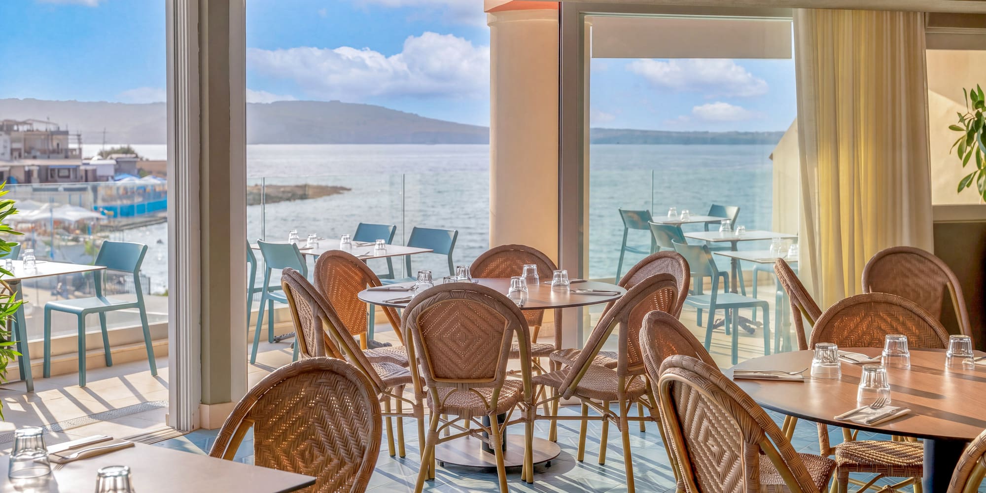 Coastal restaurant dining room with wicker chairs, set tables, and ocean view.