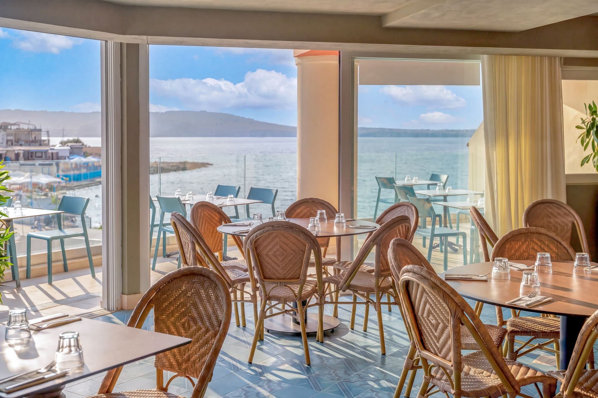 Coastal restaurant dining room with wicker chairs, set tables, and ocean view.