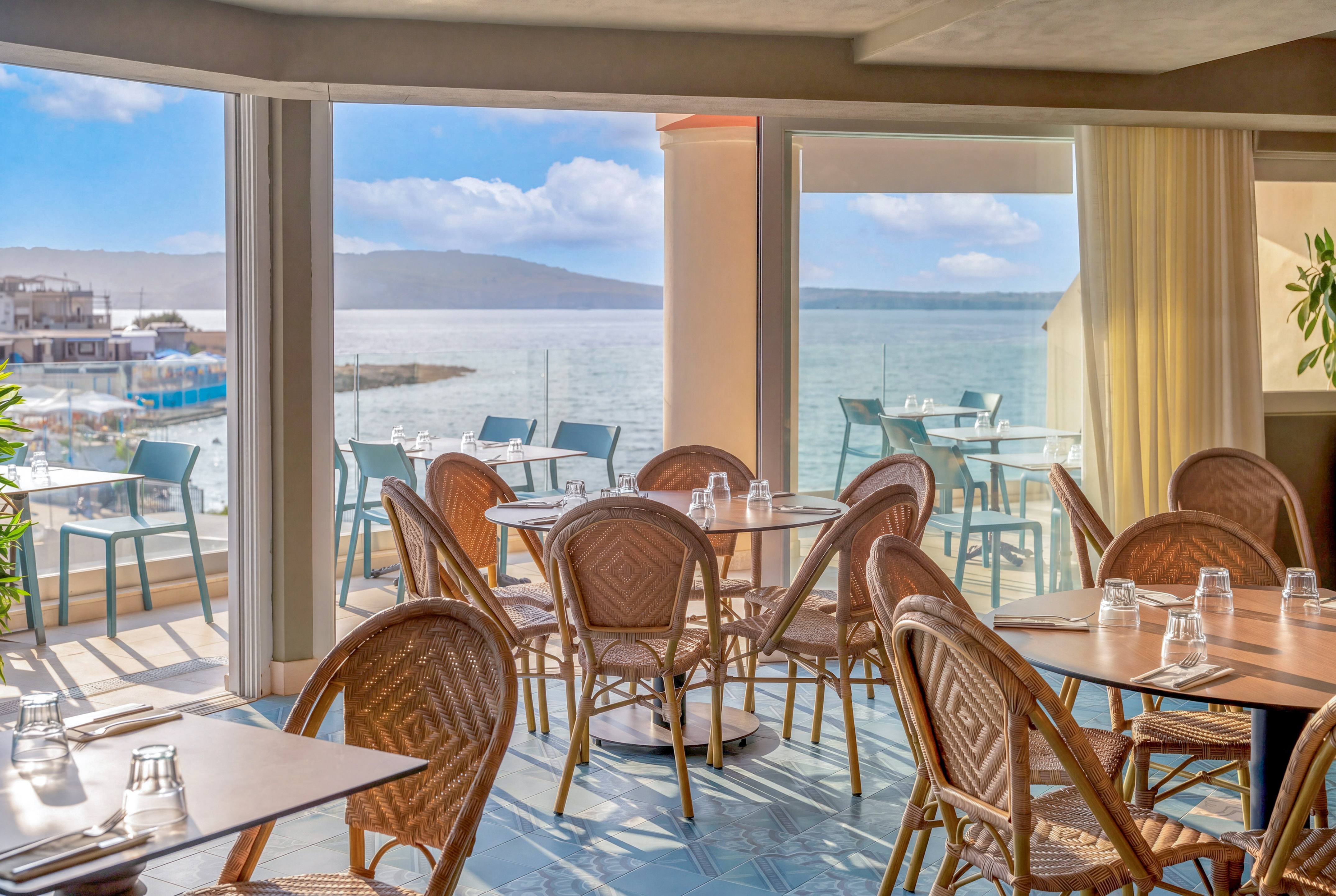 Coastal restaurant dining room with wicker chairs, set tables, and ocean view.
