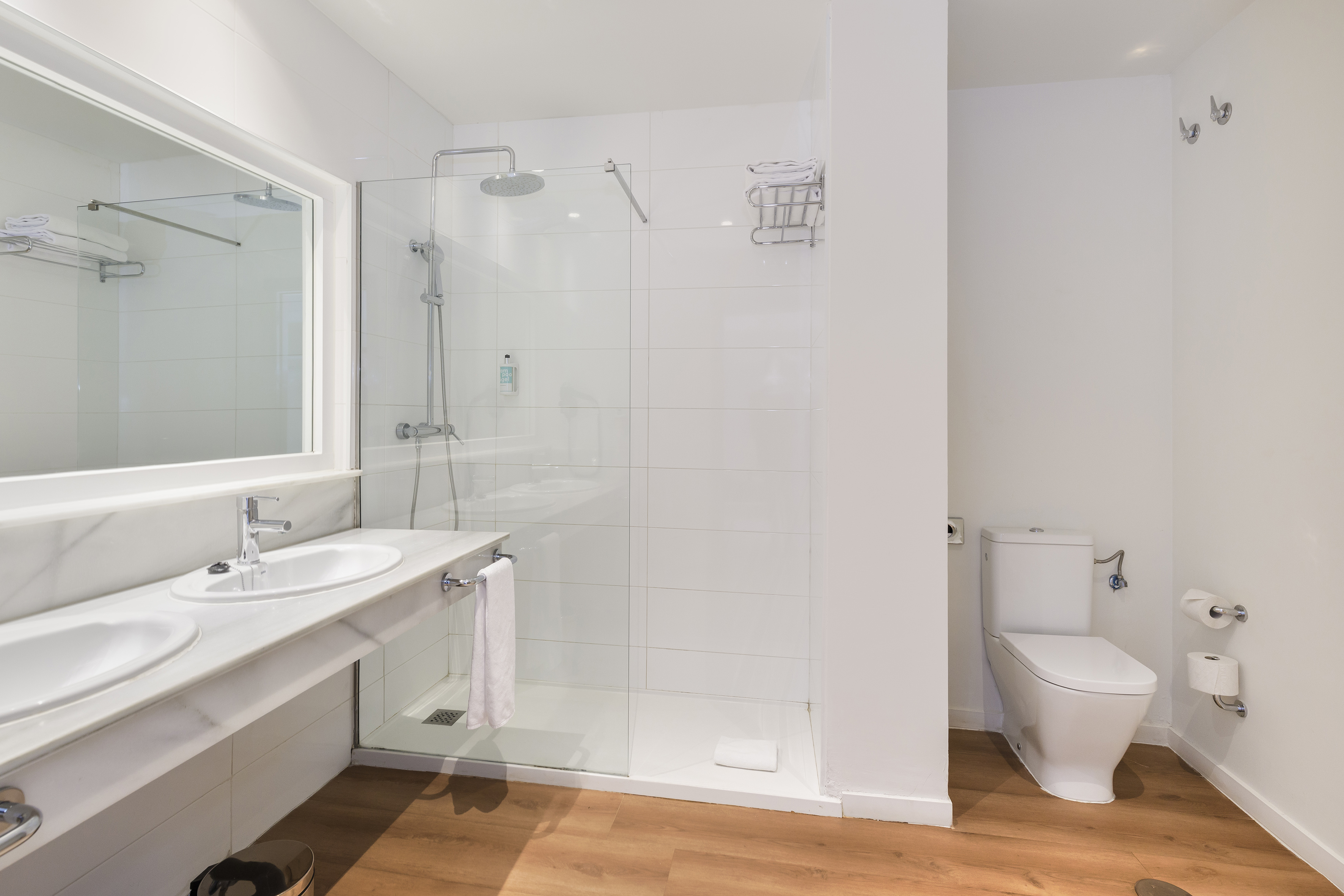 Clean white bathroom with double vanity, glass shower, toilet, and wood floor.