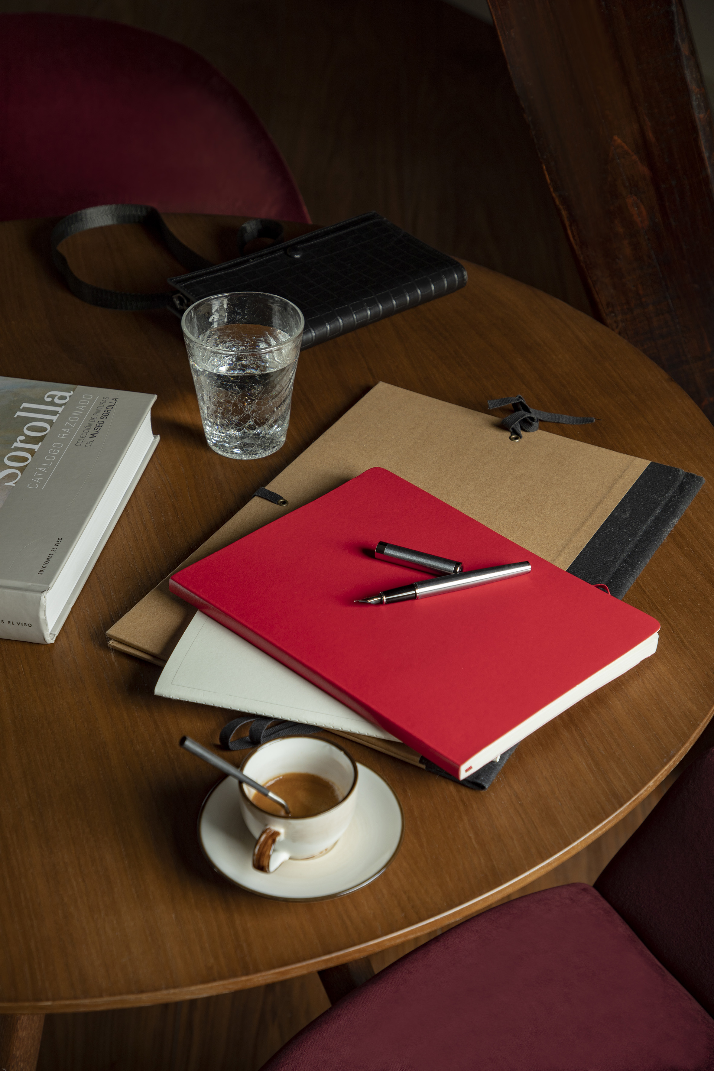 Desk setup: red notebook, pen, espresso, water glass, Sorolla book on wood.