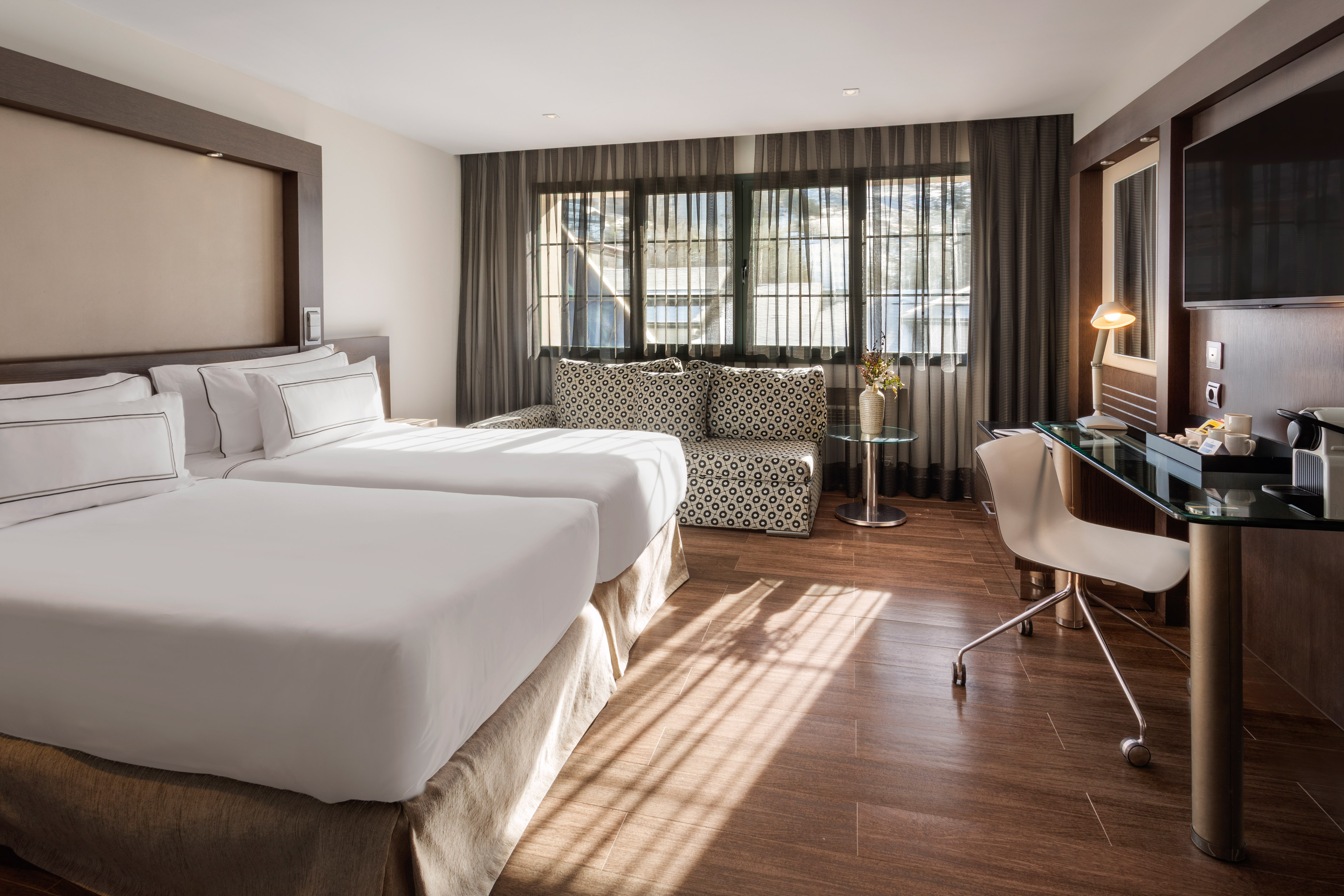 Modern hotel room with twin beds, sofa, desk, and sunlight.