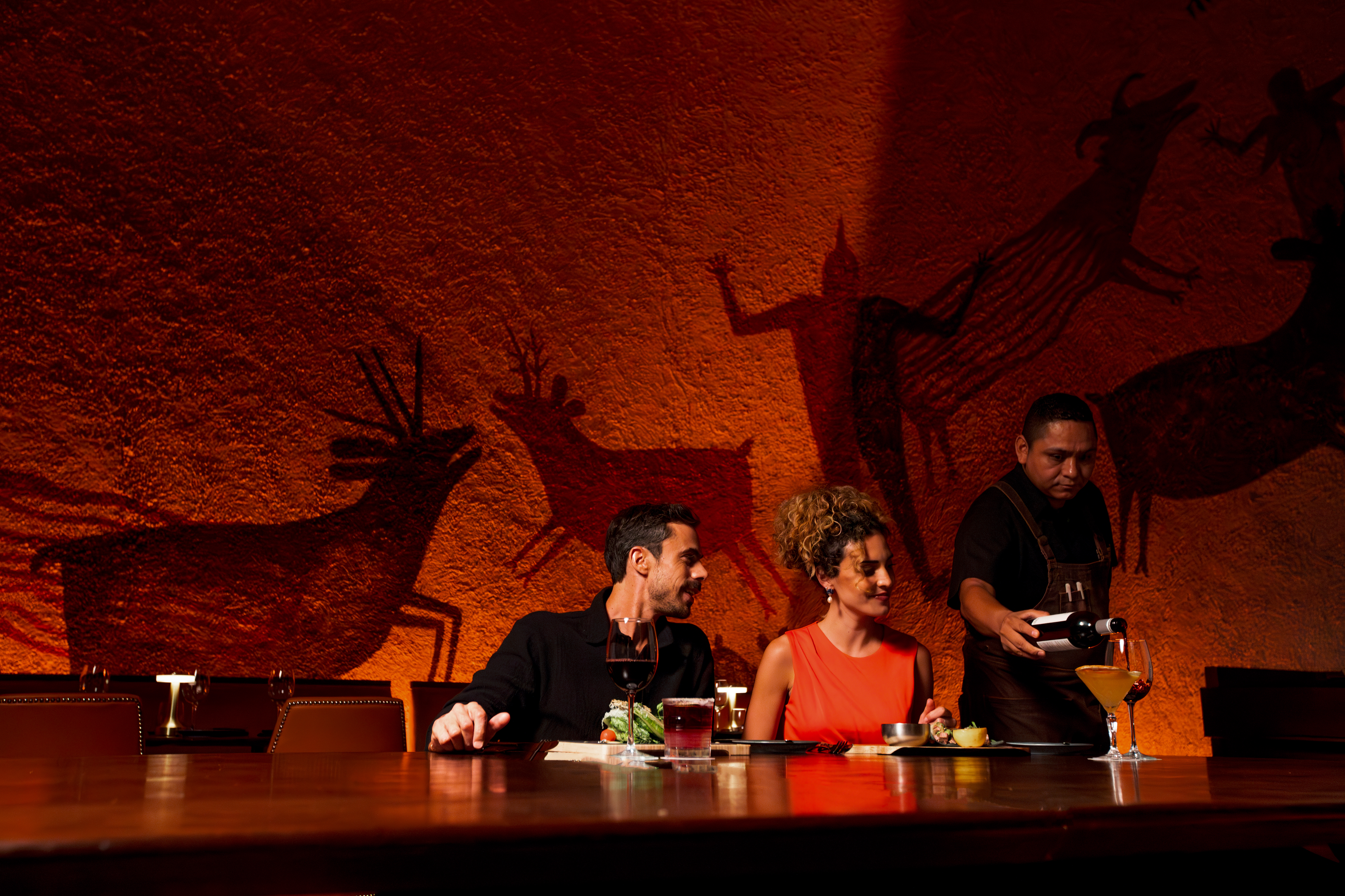 Couple dining, waiter pouring wine at restaurant with ancient cave art wall.