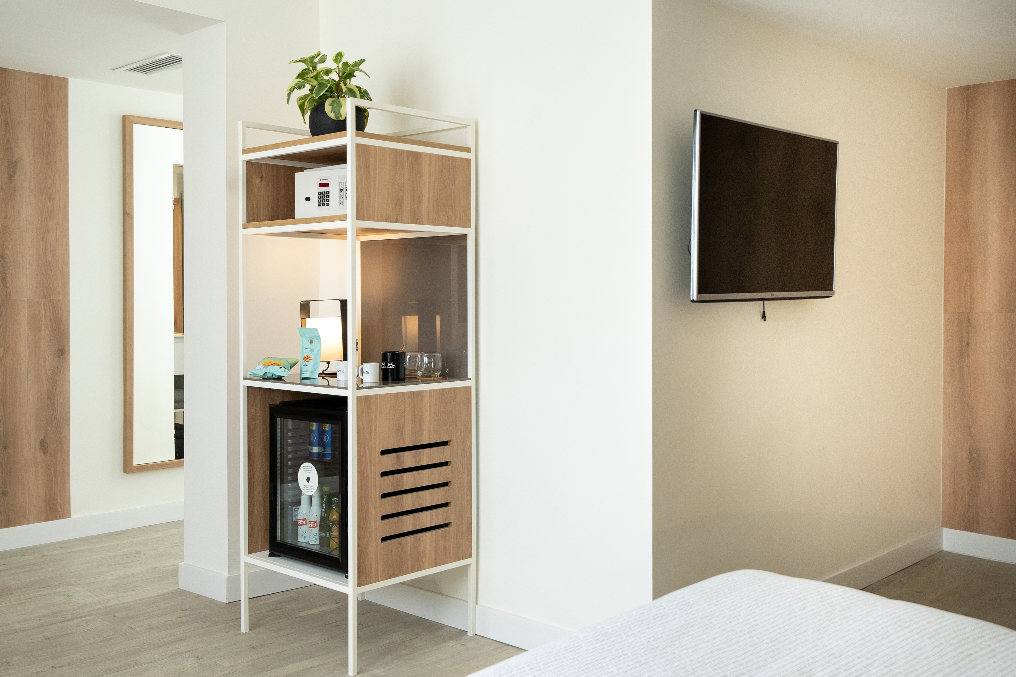 Modern hotel room with shelving unit, mini-bar, safe, TV, and mirror.