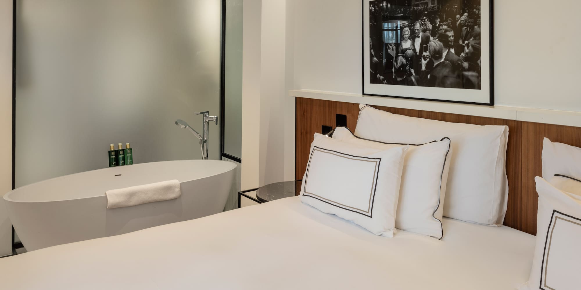 Modern hotel bedroom with white bed, freestanding bathtub, and framed art.