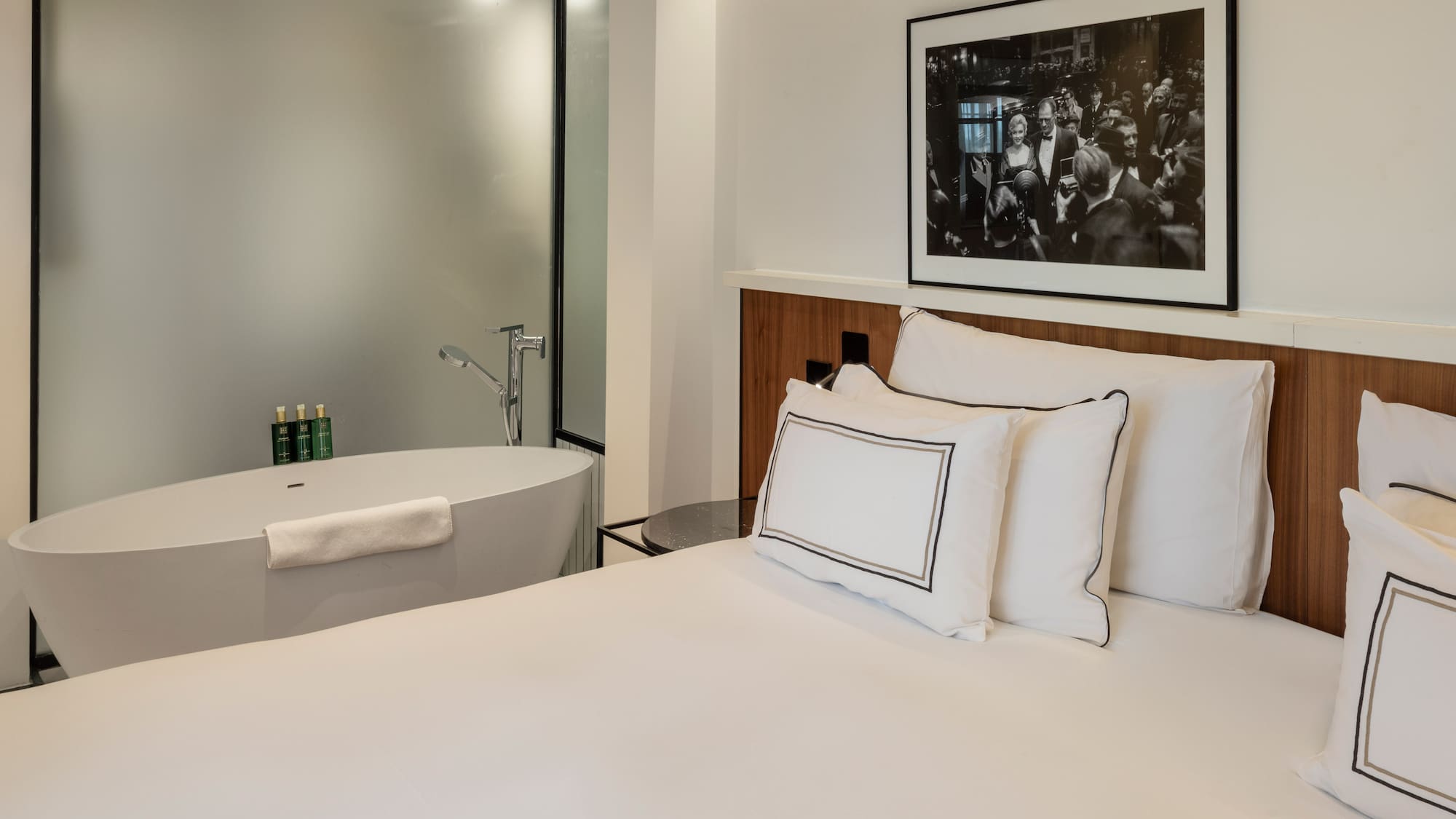 Modern hotel bedroom with white bed, freestanding bathtub, and framed art.