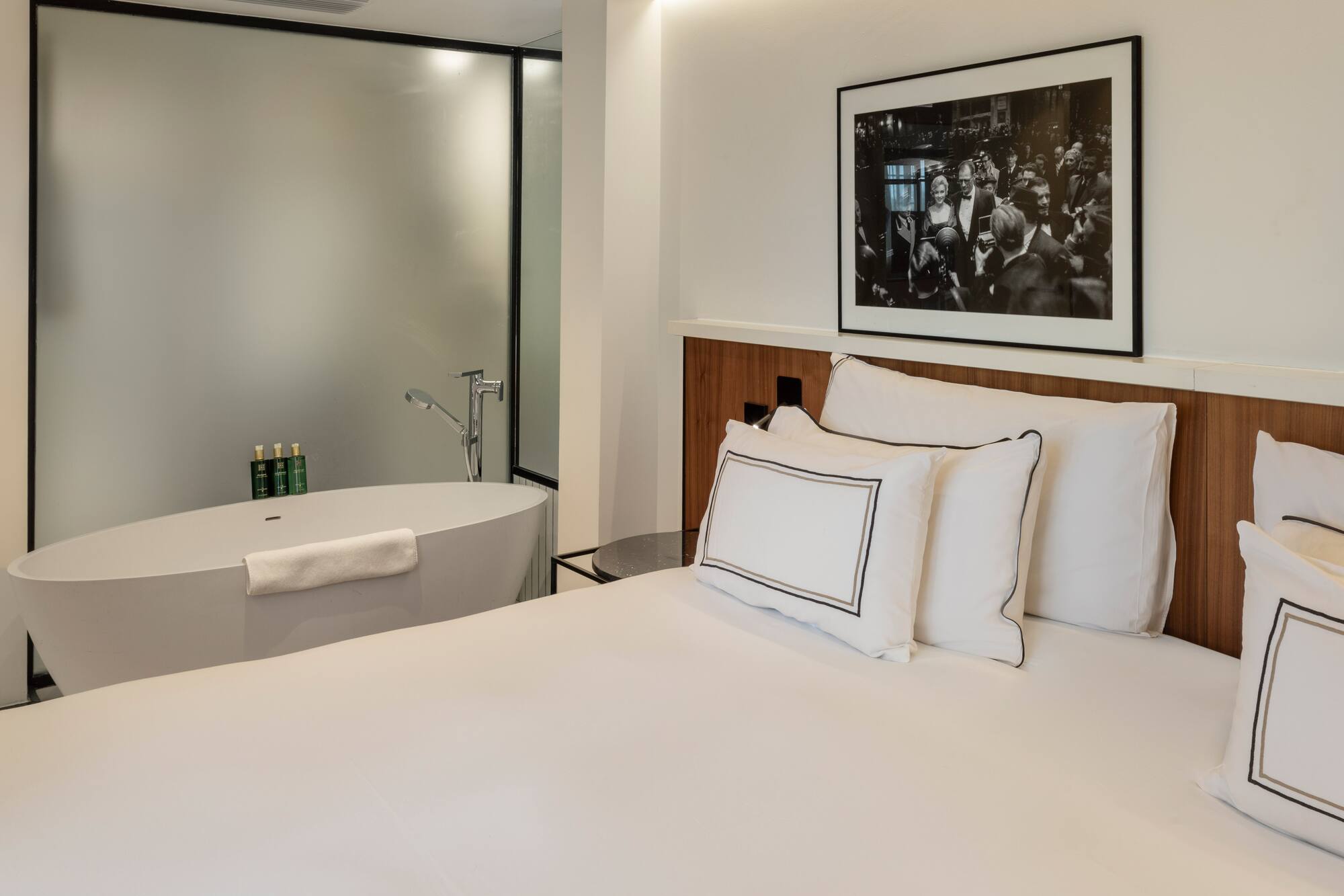 Modern hotel bedroom with white bed, freestanding bathtub, and framed art.