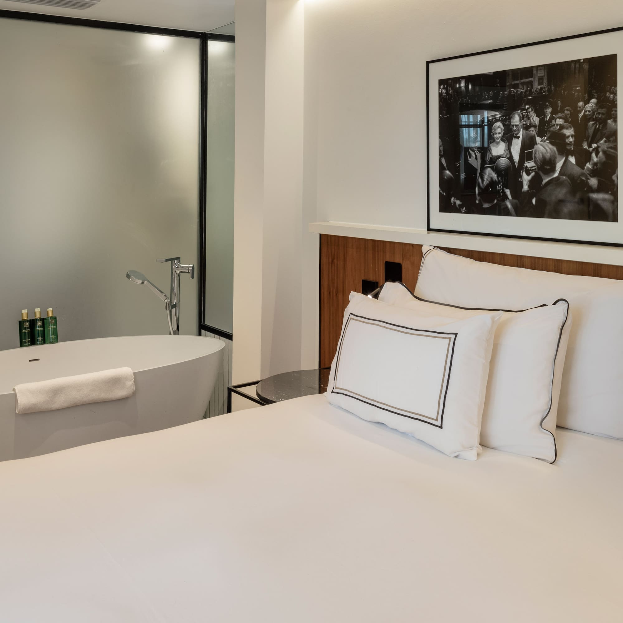 Modern hotel bedroom with white bed, freestanding bathtub, and framed art.