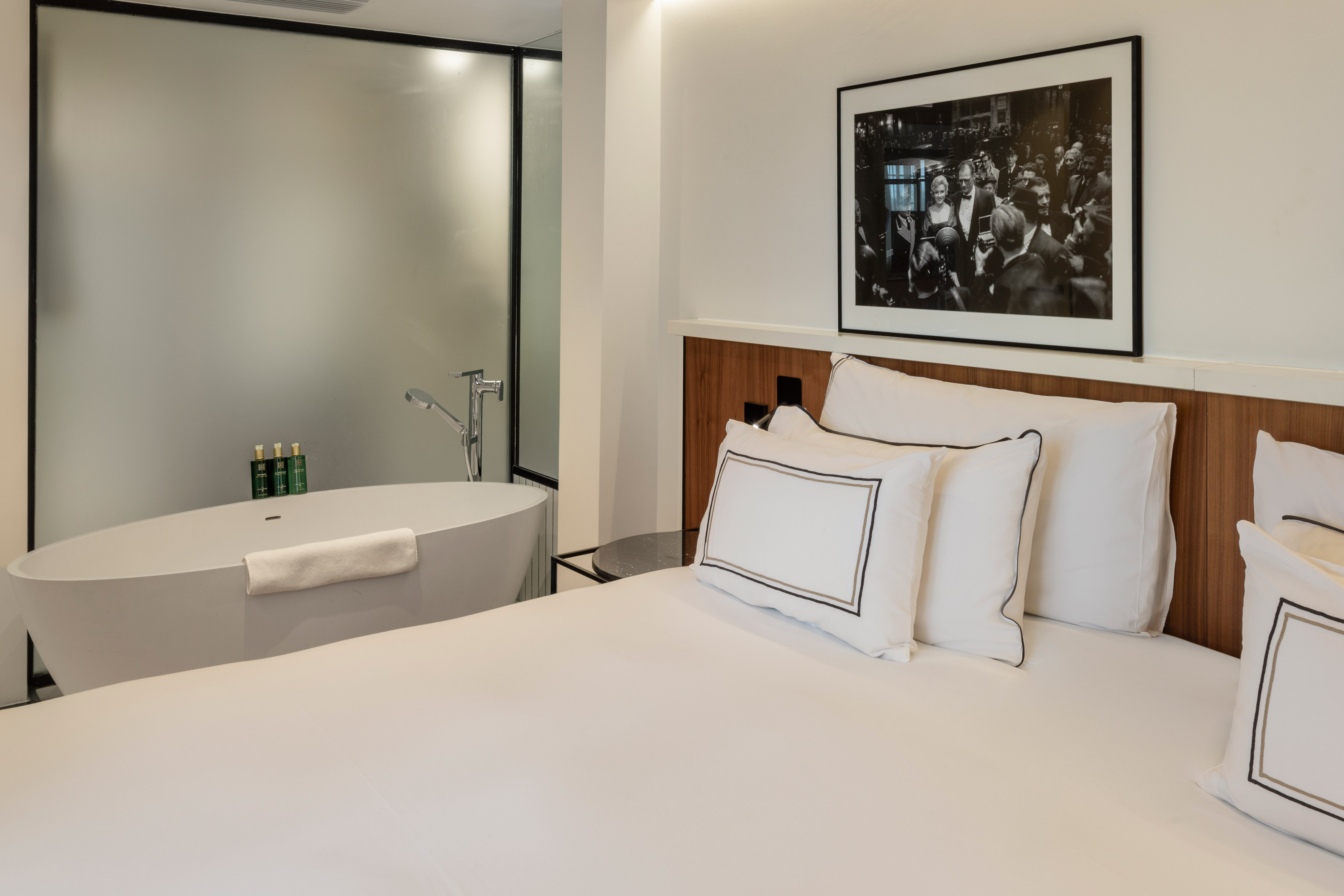 Modern hotel bedroom with white bed, freestanding bathtub, and framed art.