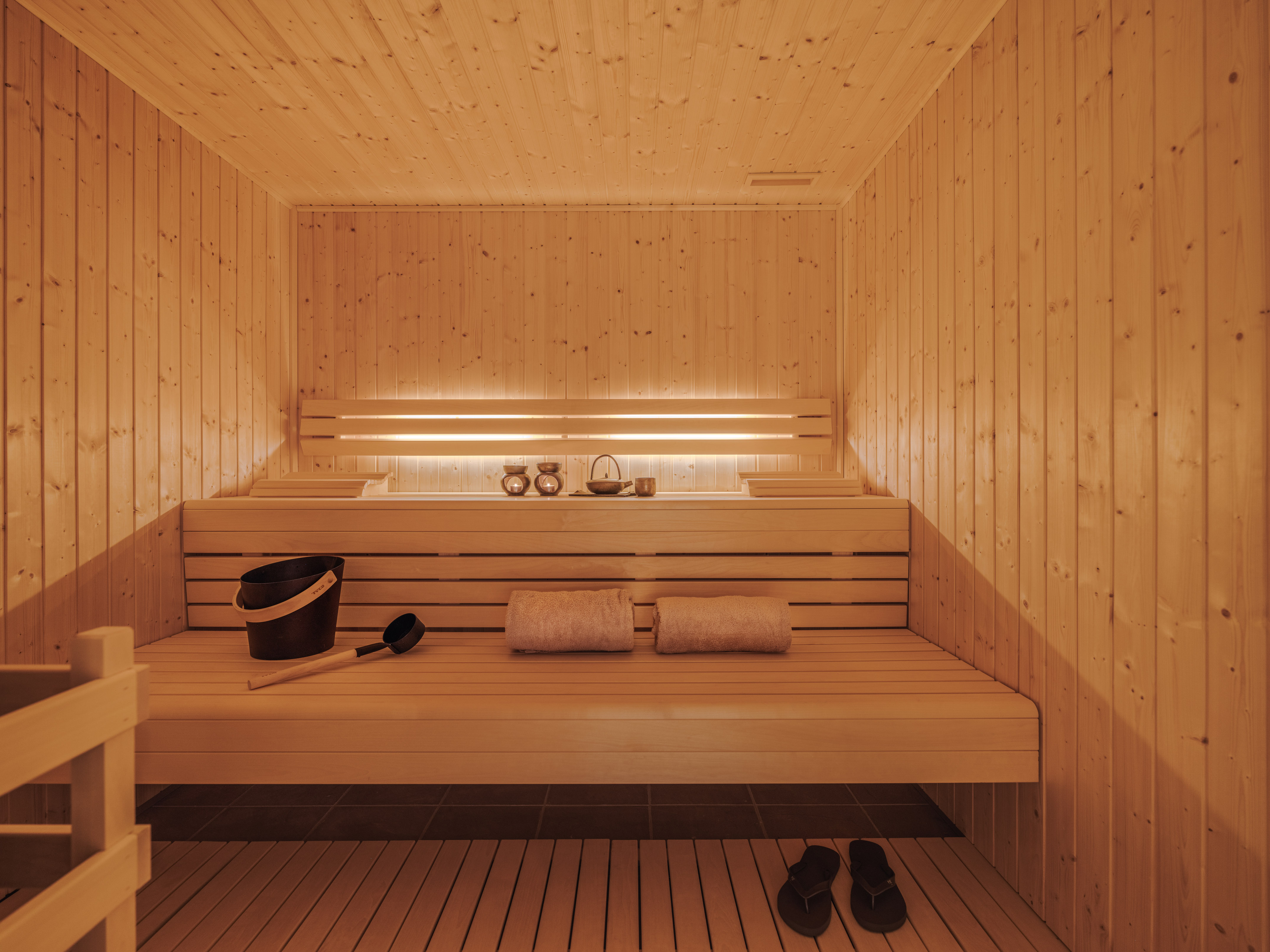 Wooden sauna with benches, towels, bucket, and warm backlighting.