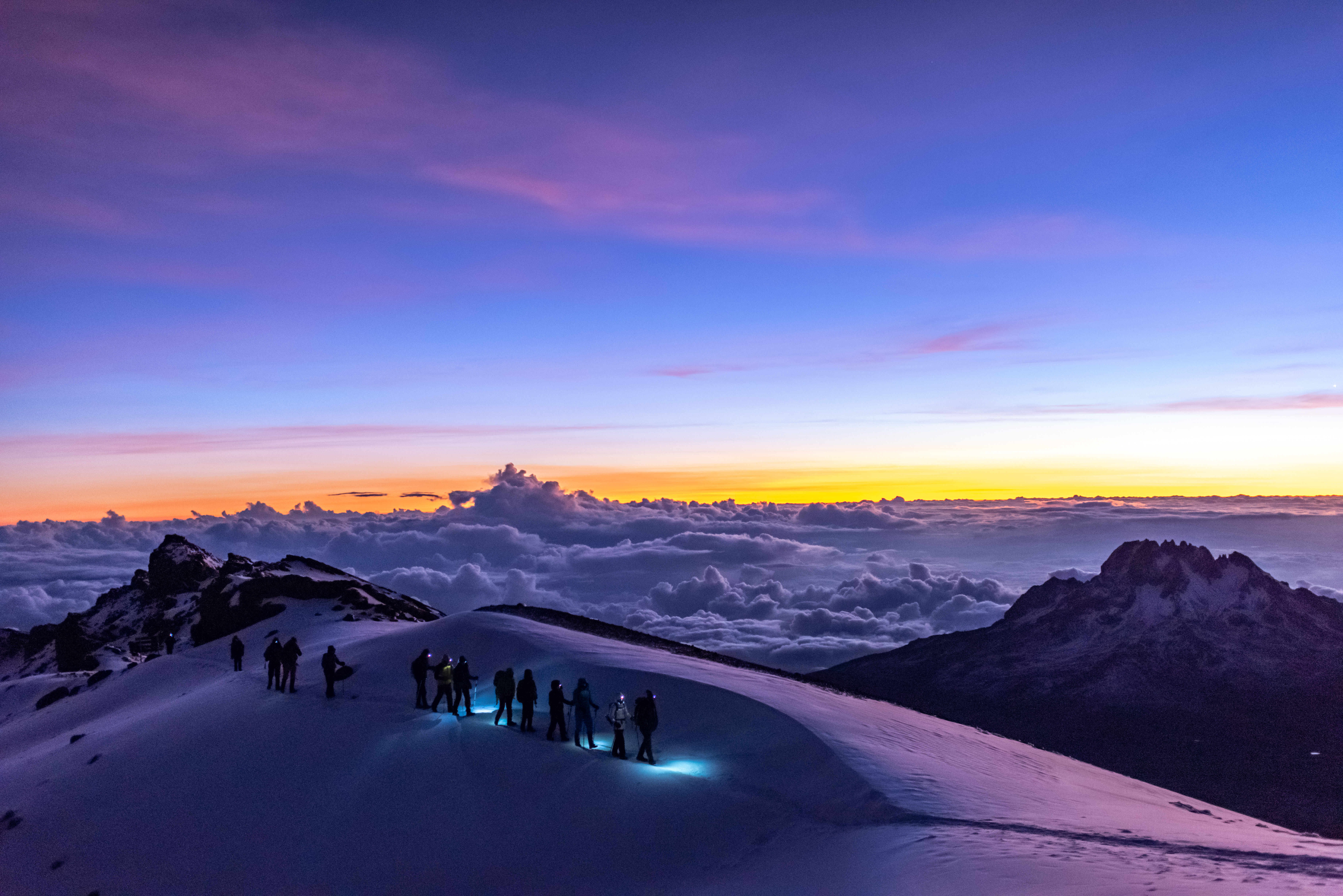 Kilimanjaro hikers with headlamps on snowy ridge at dawn, colorful sky.