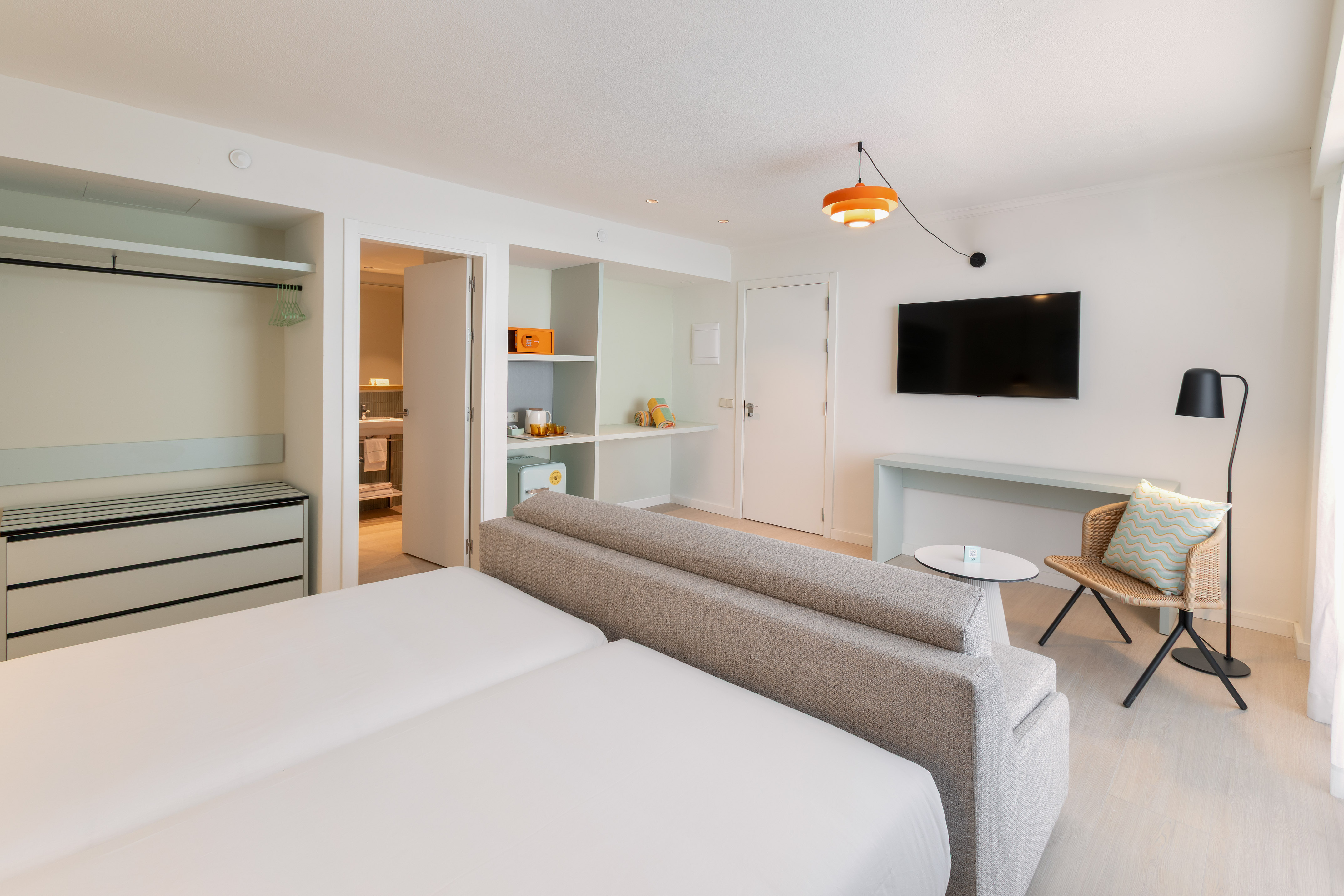 Modern hotel suite with twin beds, sofa, TV, open wardrobe, and desk area.