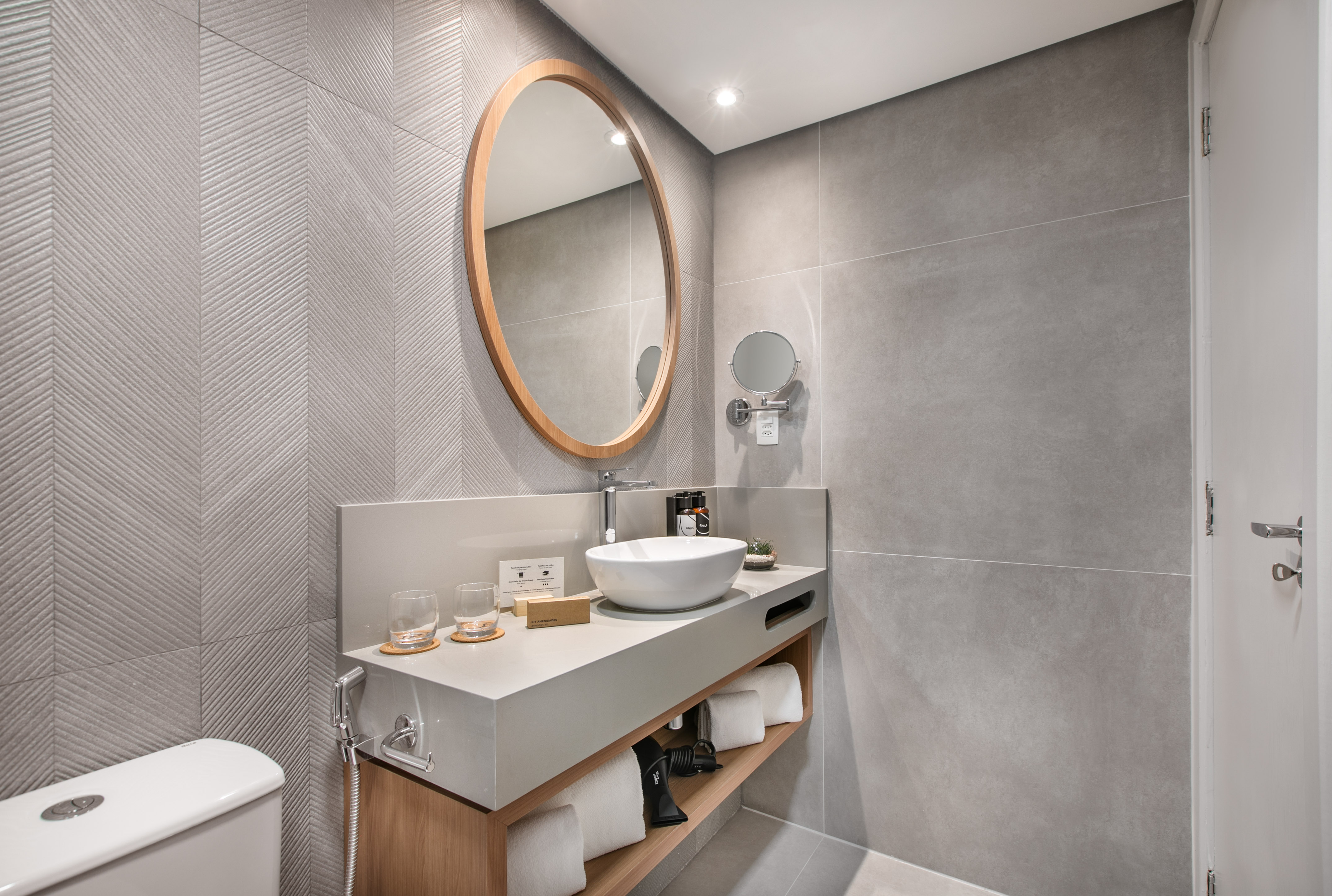 Modern hotel bathroom vanity with oval mirror, white vessel sink, and gray tiles.