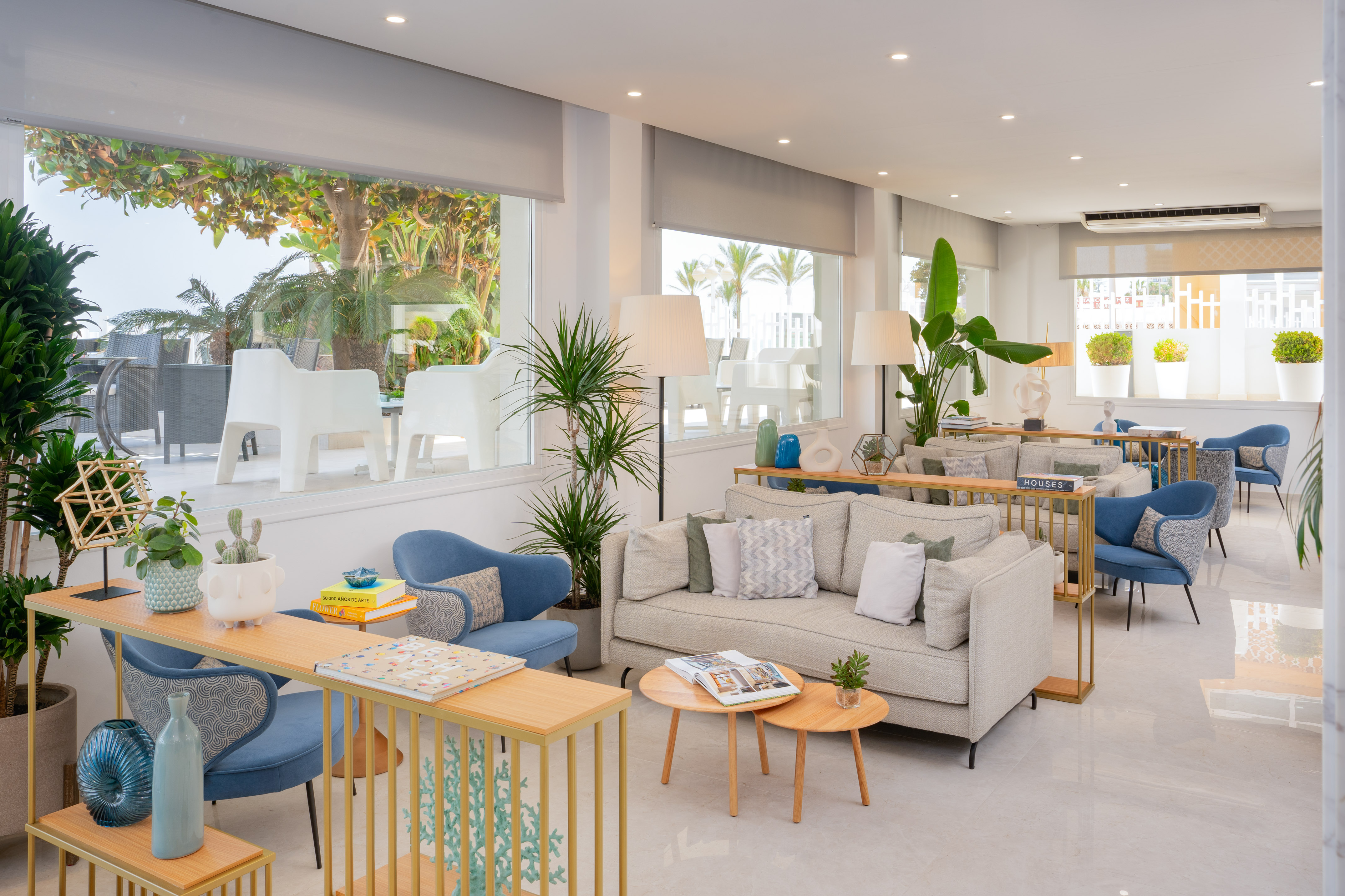 Modern hotel lobby with beige sofas, blue armchairs, and large windows overlooking tropical plants.