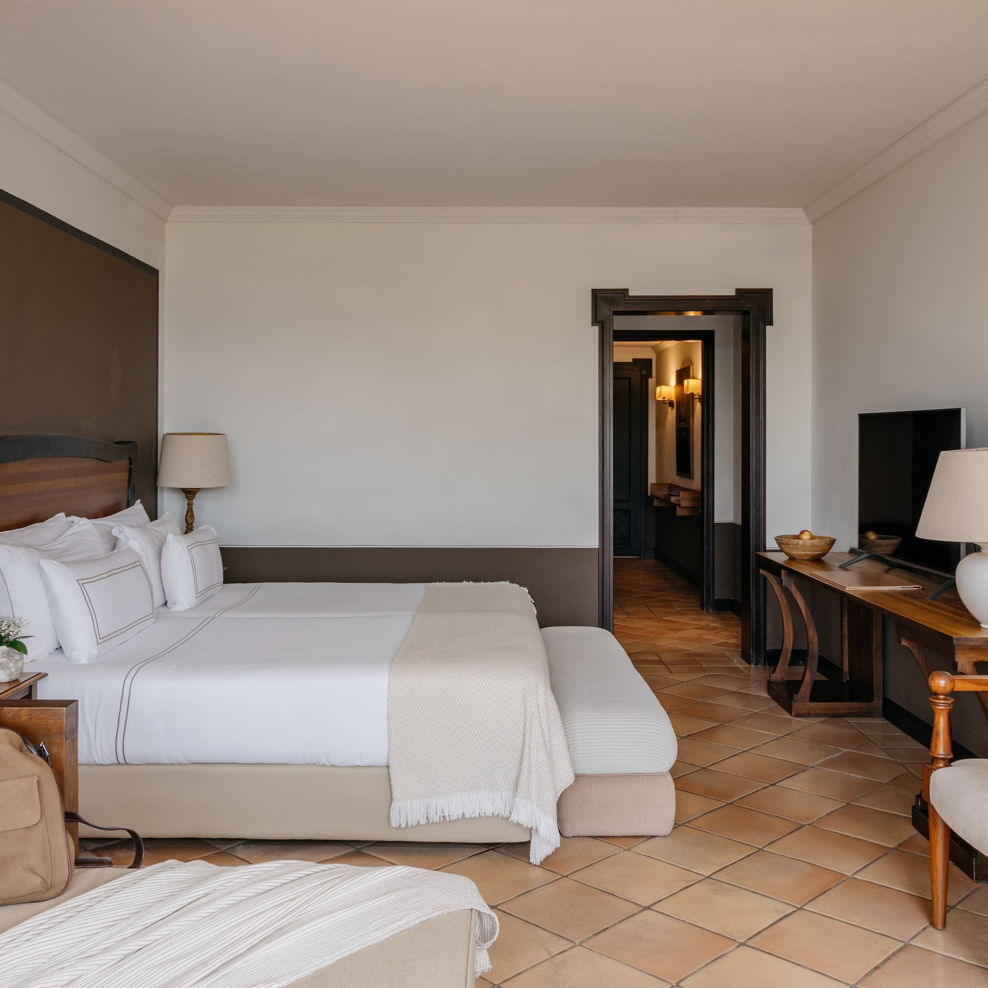 Luxury hotel bedroom with king bed, white linens, wood furniture, terracotta floor.
