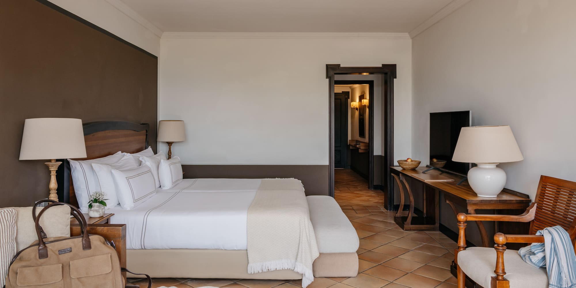 Luxury hotel bedroom with king bed, white linens, wood furniture, terracotta floor.