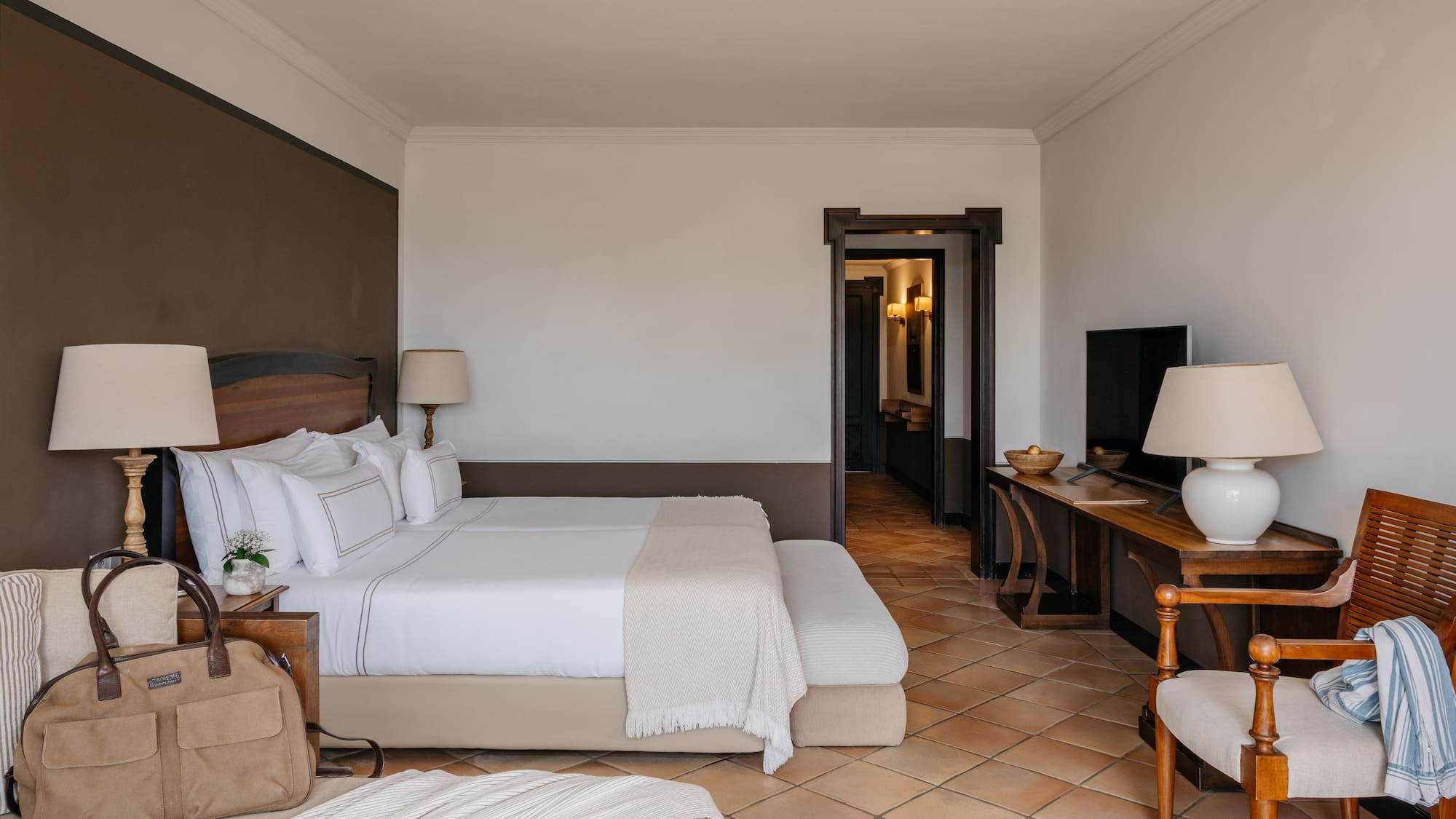 Luxury hotel bedroom with king bed, white linens, wood furniture, terracotta floor.