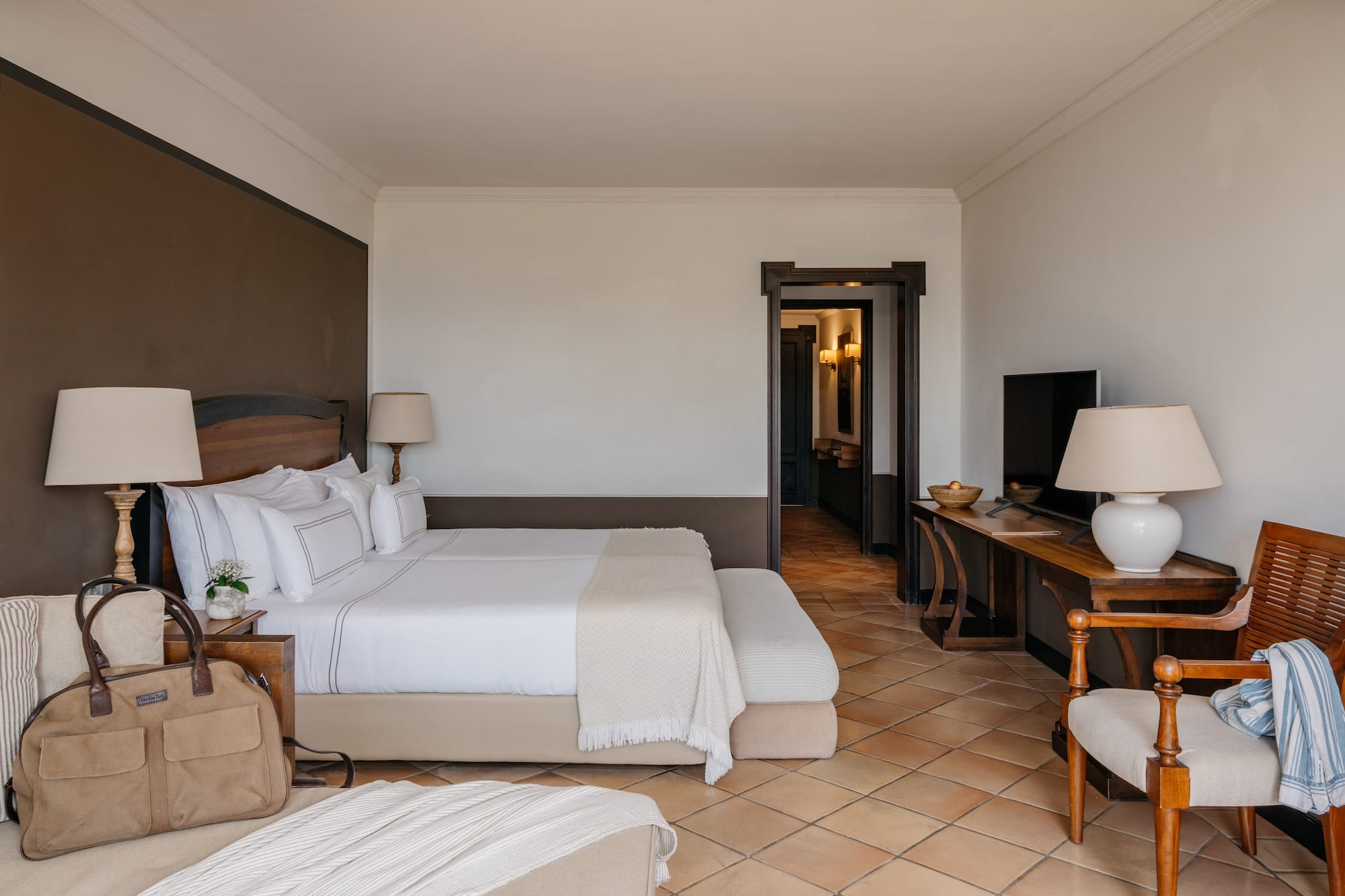 Luxury hotel bedroom with king bed, white linens, wood furniture, terracotta floor.